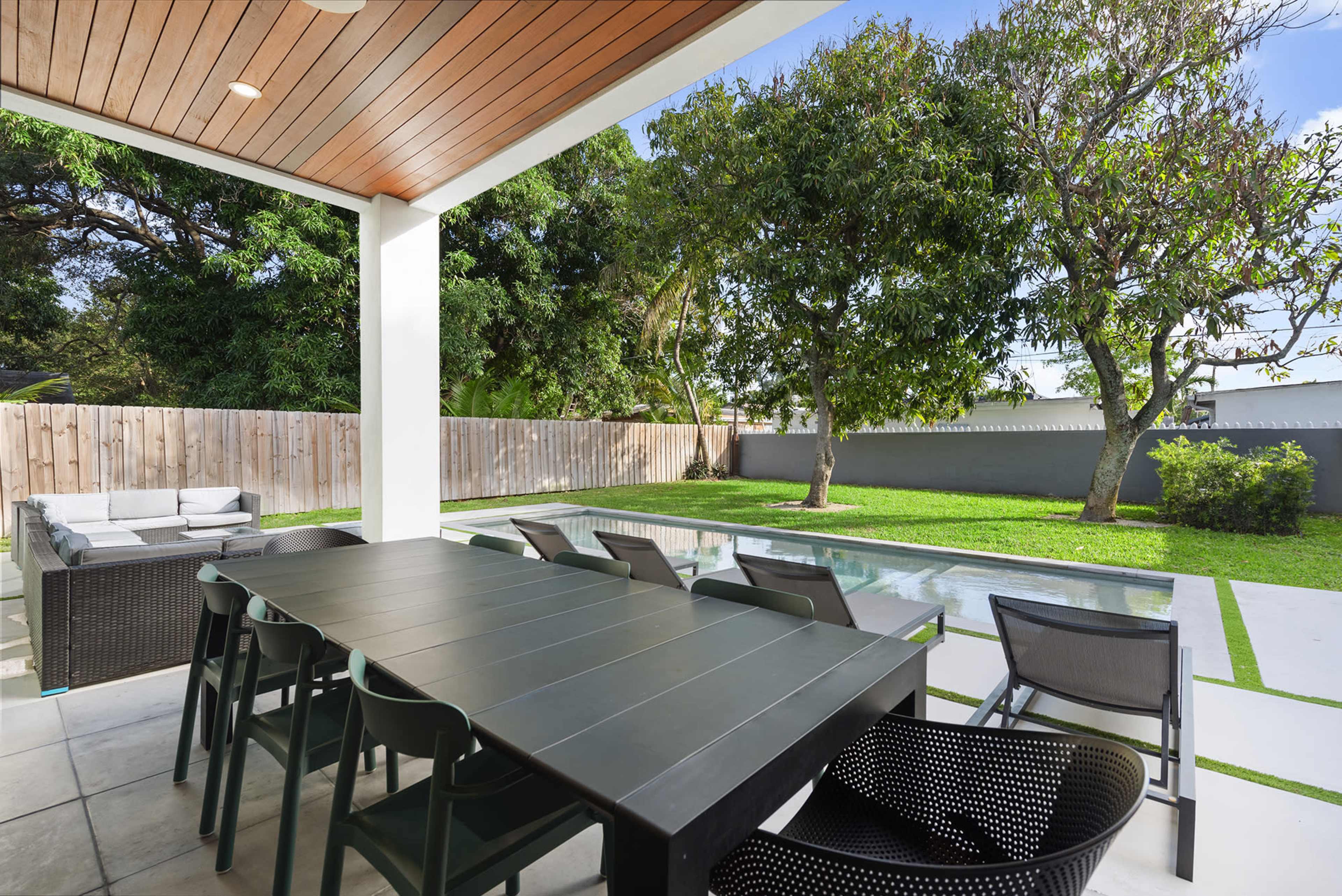 The image shows an outdoor dining area with a large black table and chairs, overlooking a lawn and swimming pool surrounded by trees.