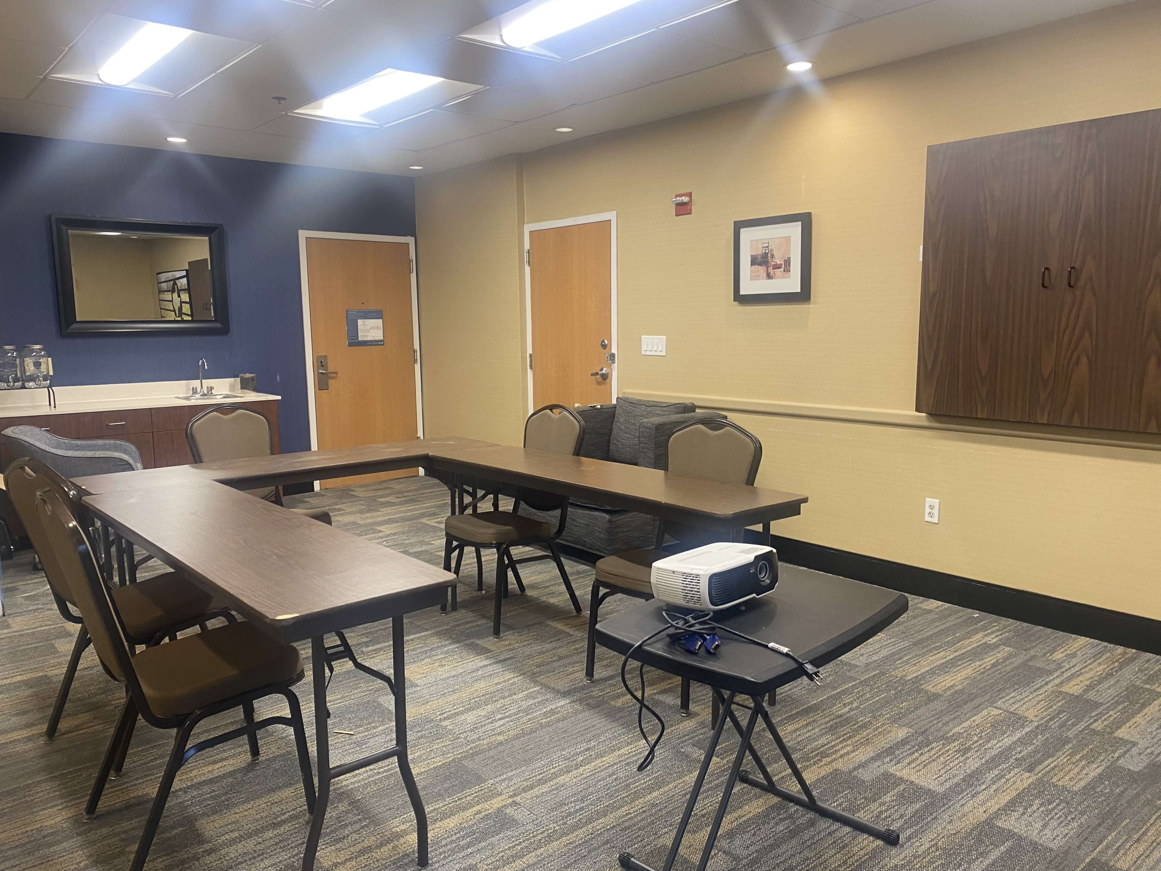 A meeting room with a U-shaped arrangement of tables and chairs, a projector on a small table, and a kitchenette area in the background.