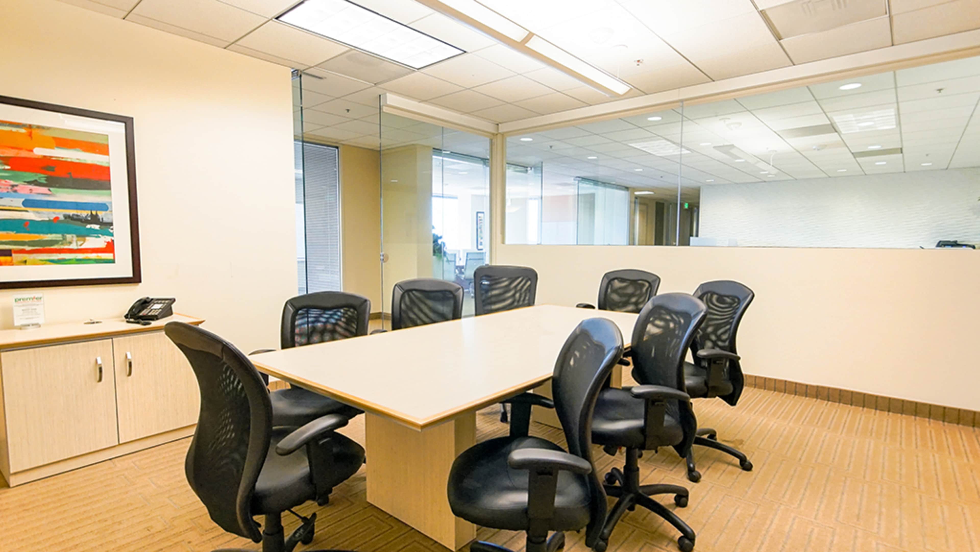 A conference room features a large table surrounded by black swivel chairs, with a glass partition and modern office decor in the background.