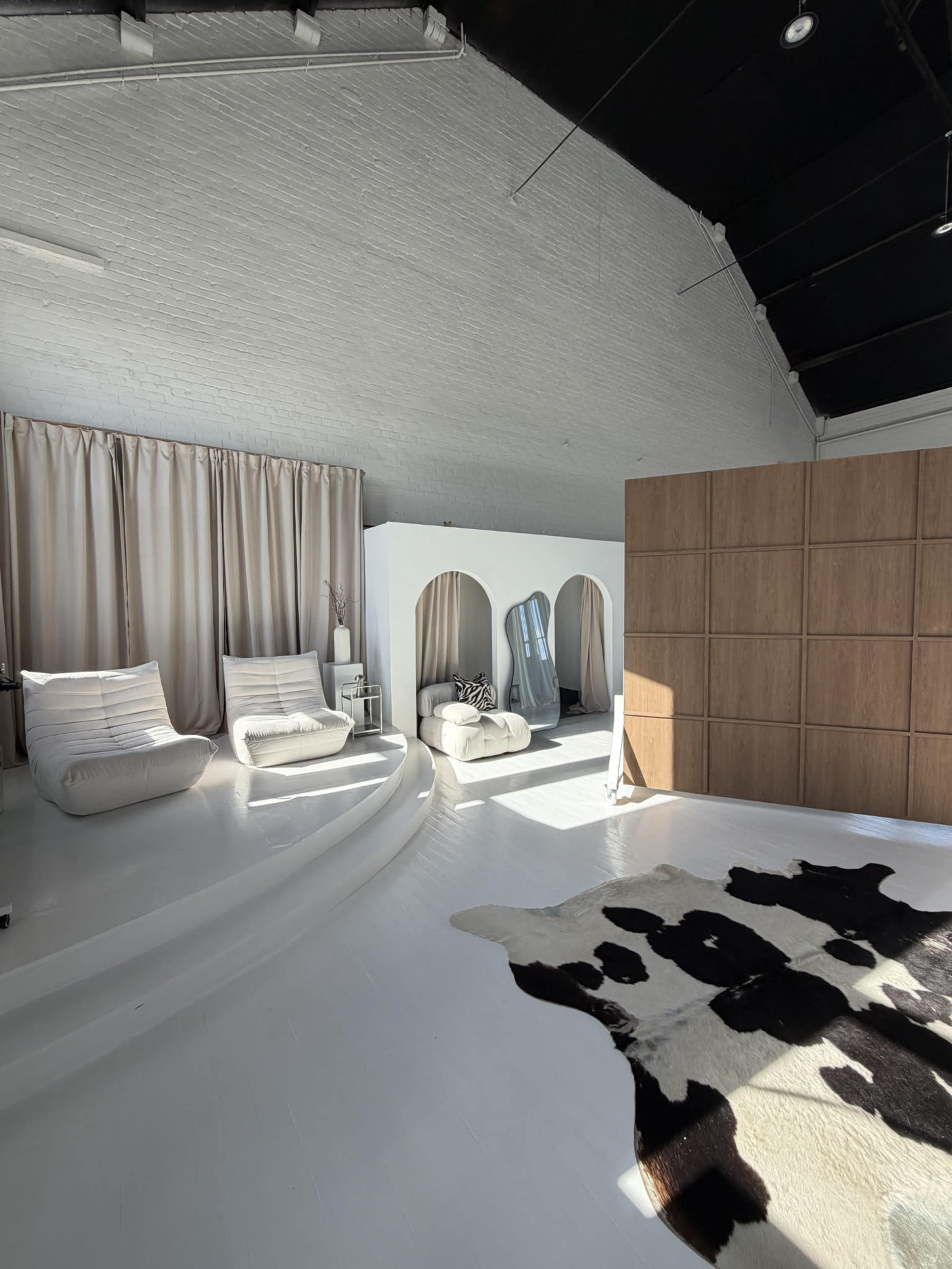 The image shows a spacious, bright interior featuring white walls, two curved chairs, a cowhide rug on the floor, and sections divided by wooden panels and curtains.
