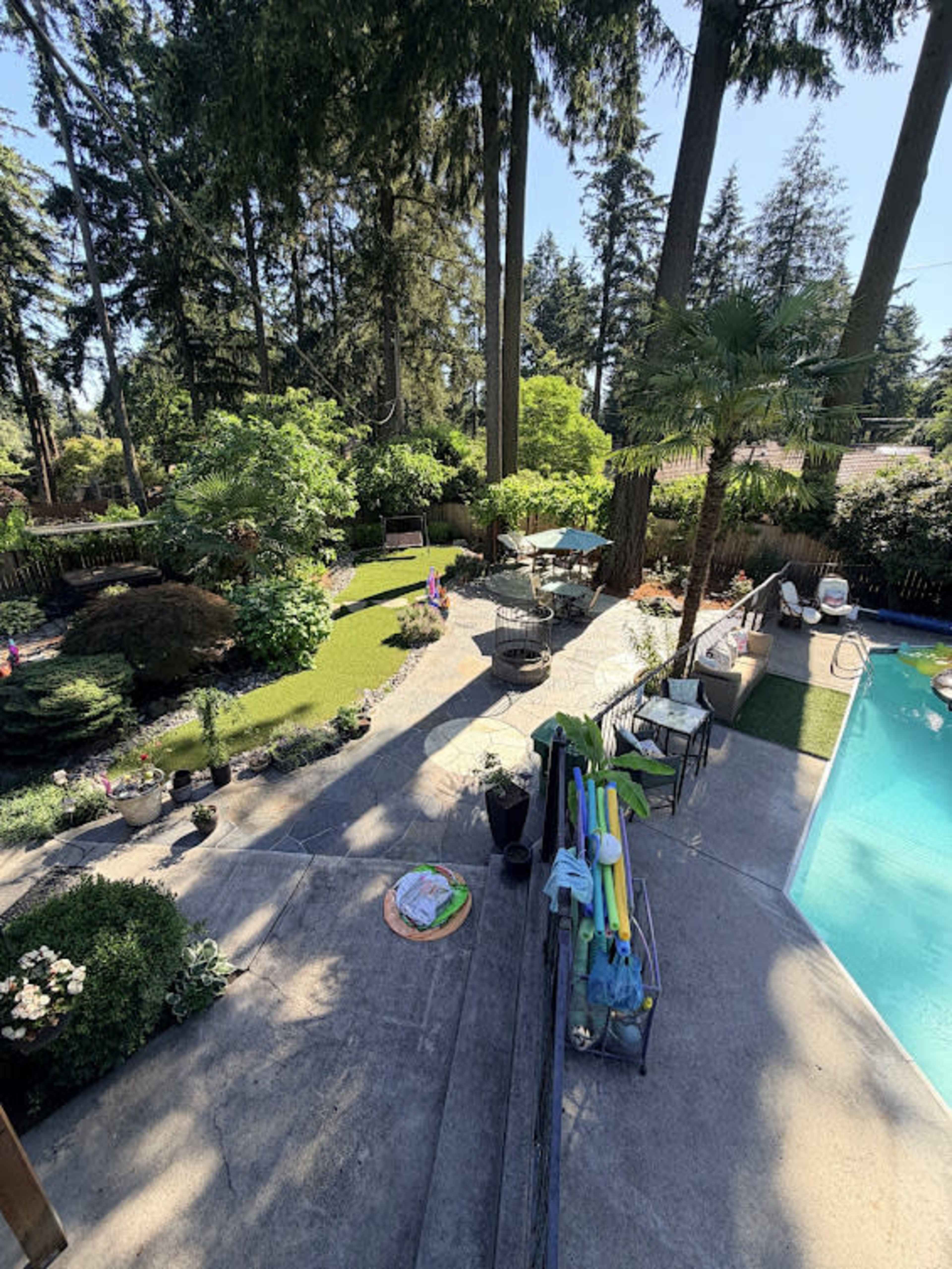 The image shows a backyard with a swimming pool, landscaped garden, patio furniture, and trees under a blue sky.