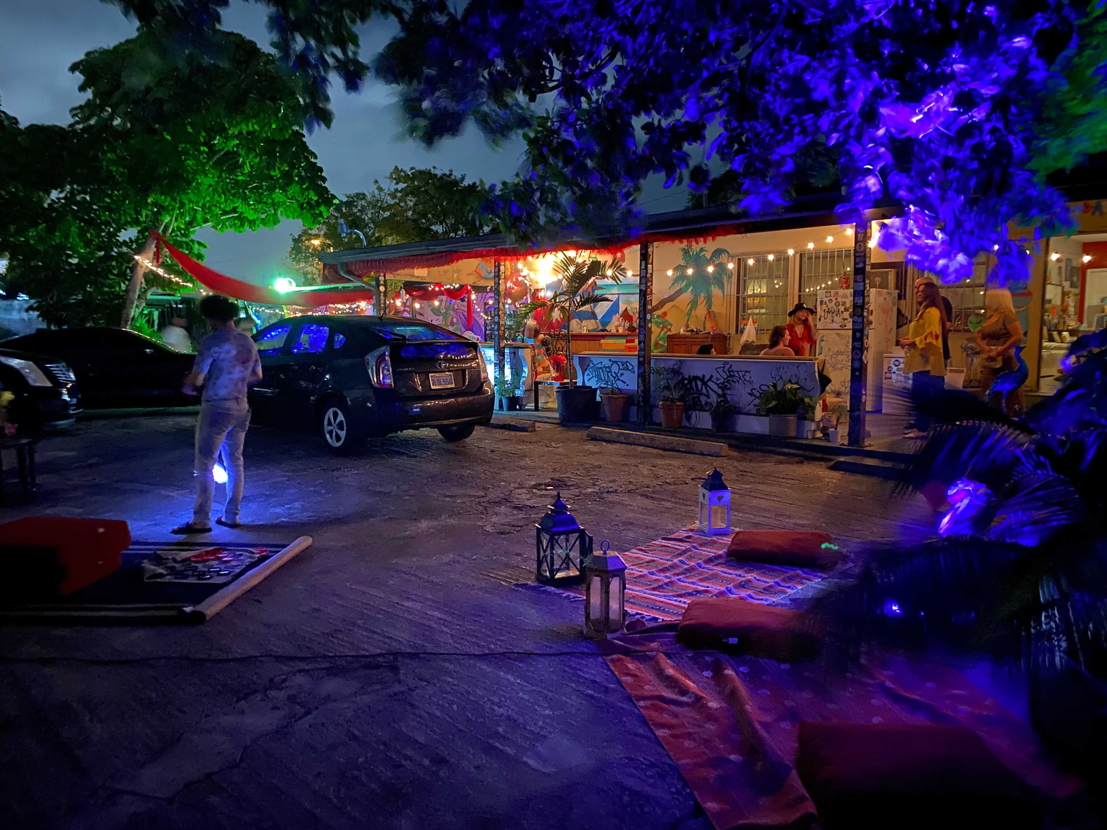 A colorful outdoor gathering features guests socializing near a brightly lit restaurant, with a parked car and decorative lanterns on the ground.