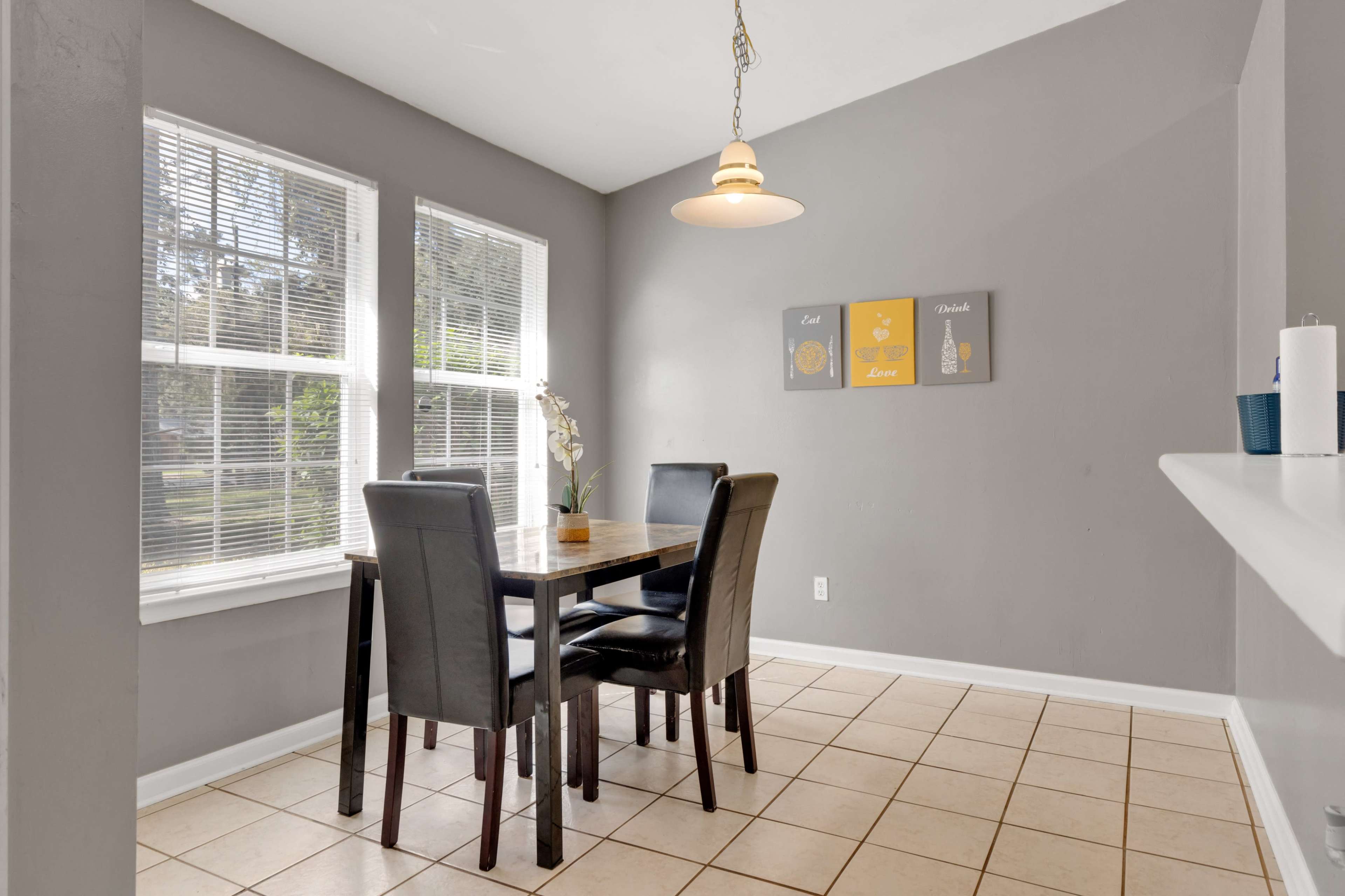 A dining area features a wooden table surrounded by black chairs, with two windows allowing natural light and gray walls decorated with framed artwork.