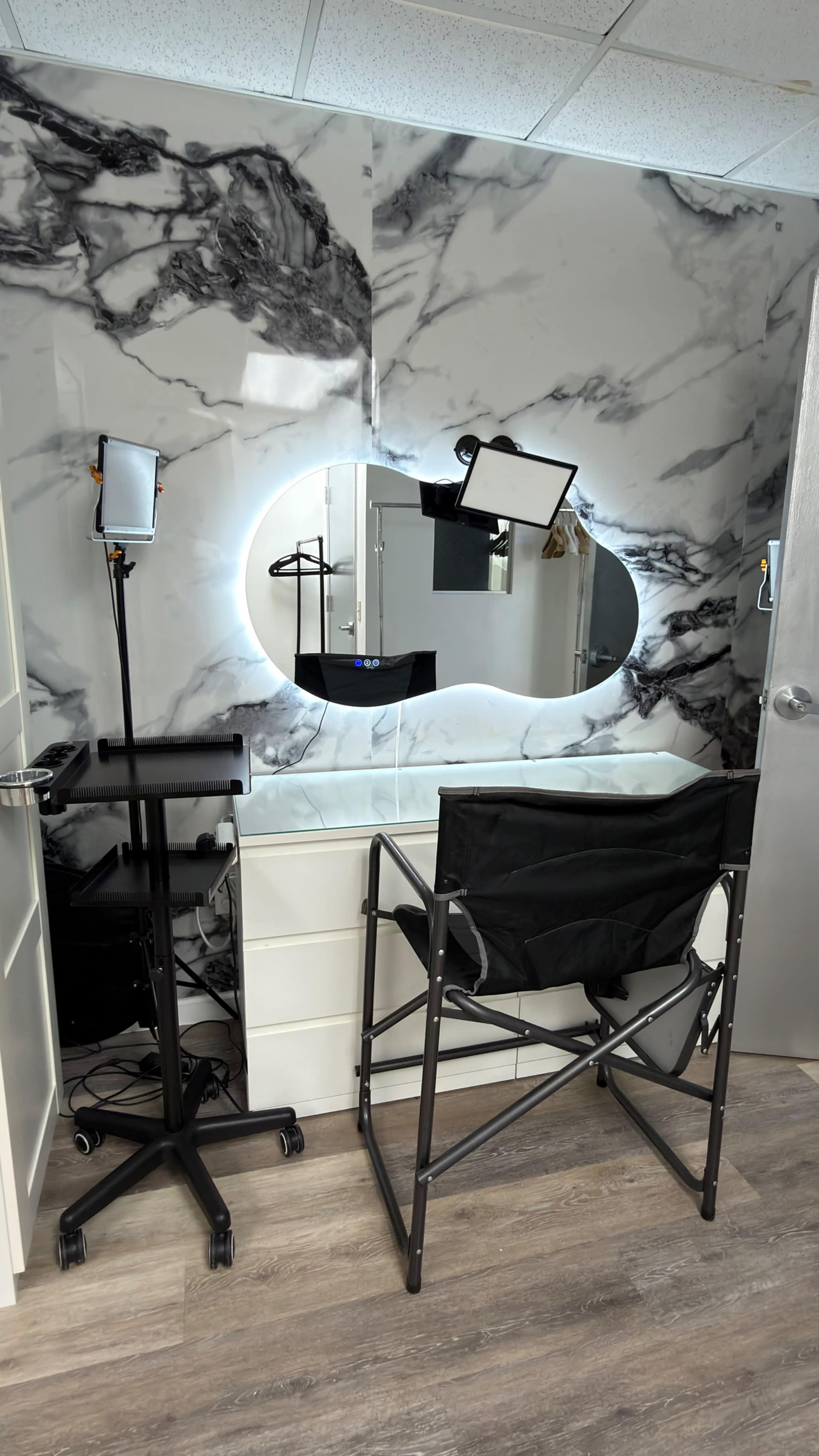 The image shows a makeup station featuring a large oval mirror with backlighting, a director's chair, and a portable lighting setup against a marble-patterned wall.