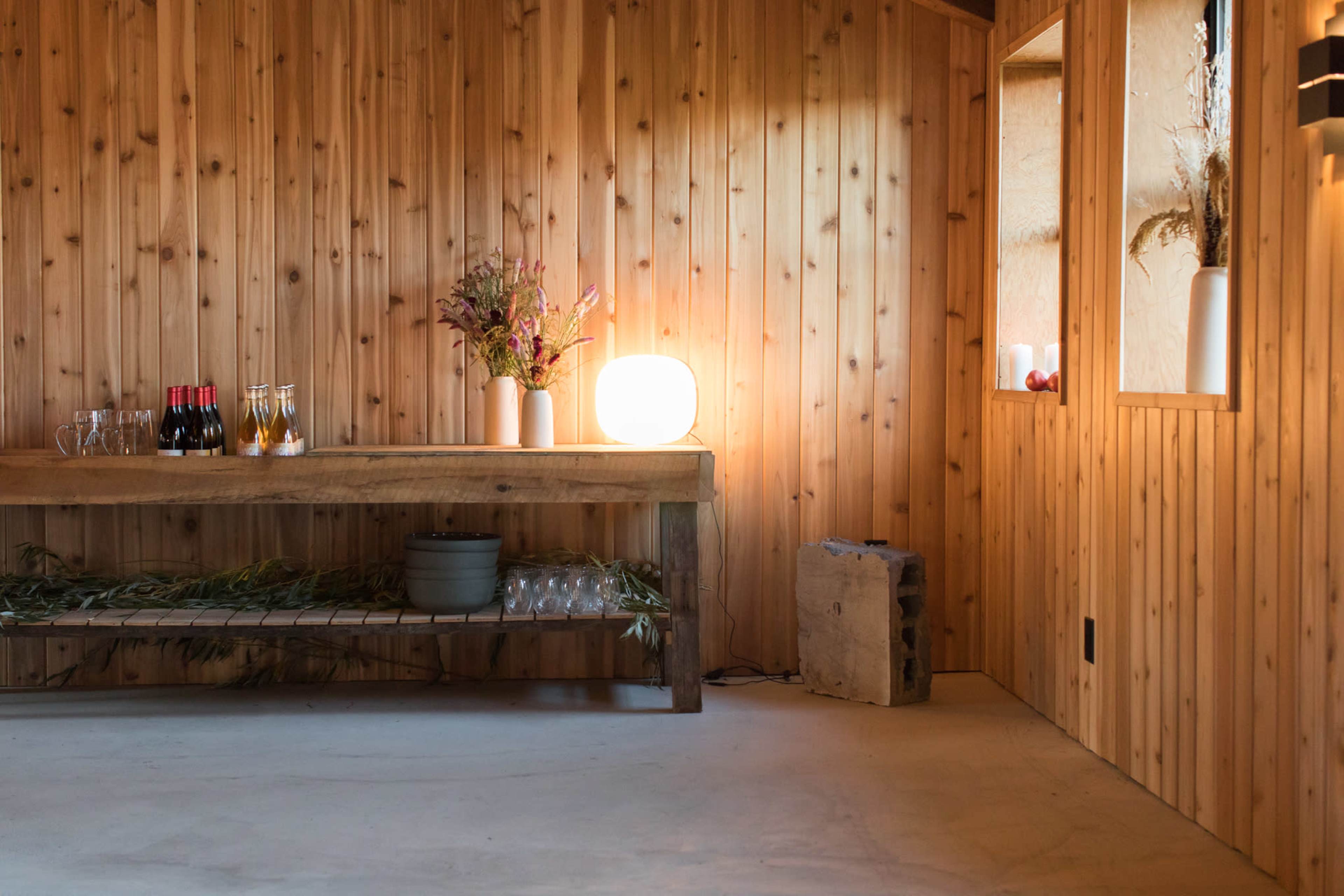 A wooden room features a table with bottles, glassware, and flowers, illuminated by a round light on the floor.
