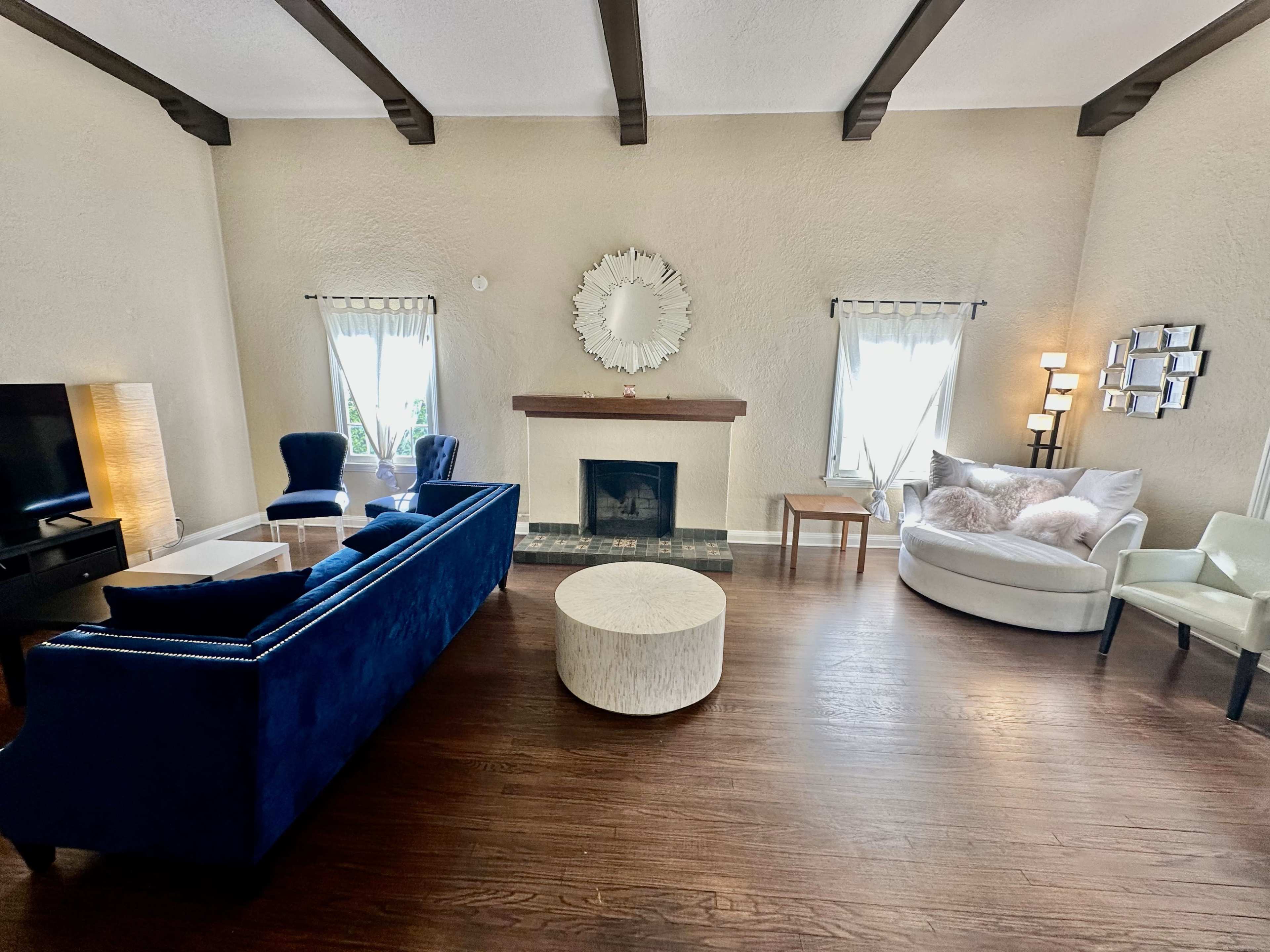 The image shows a spacious living room with a blue sofa, a round coffee table, an armchair, and a fireplace, illuminated by natural light from two windows.