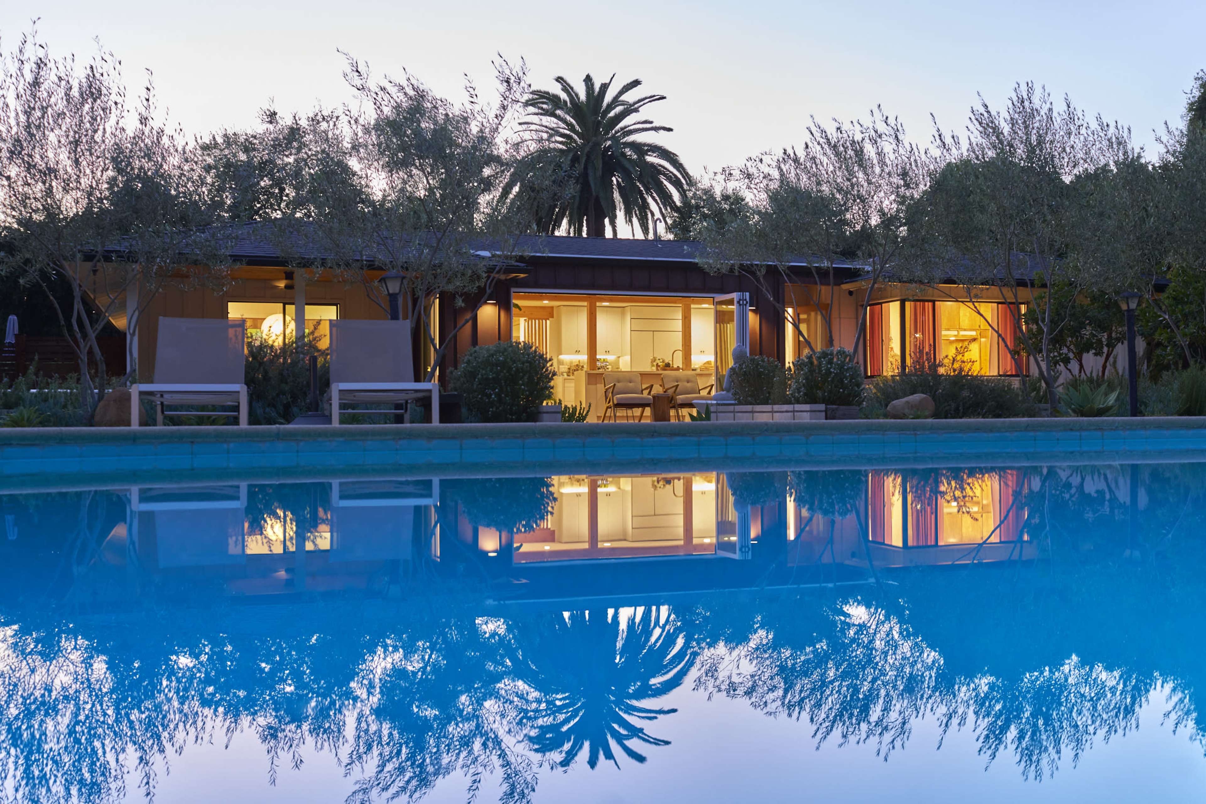 A modern home with large windows overlooks a still pool reflecting the surrounding palm trees and evening sky.
