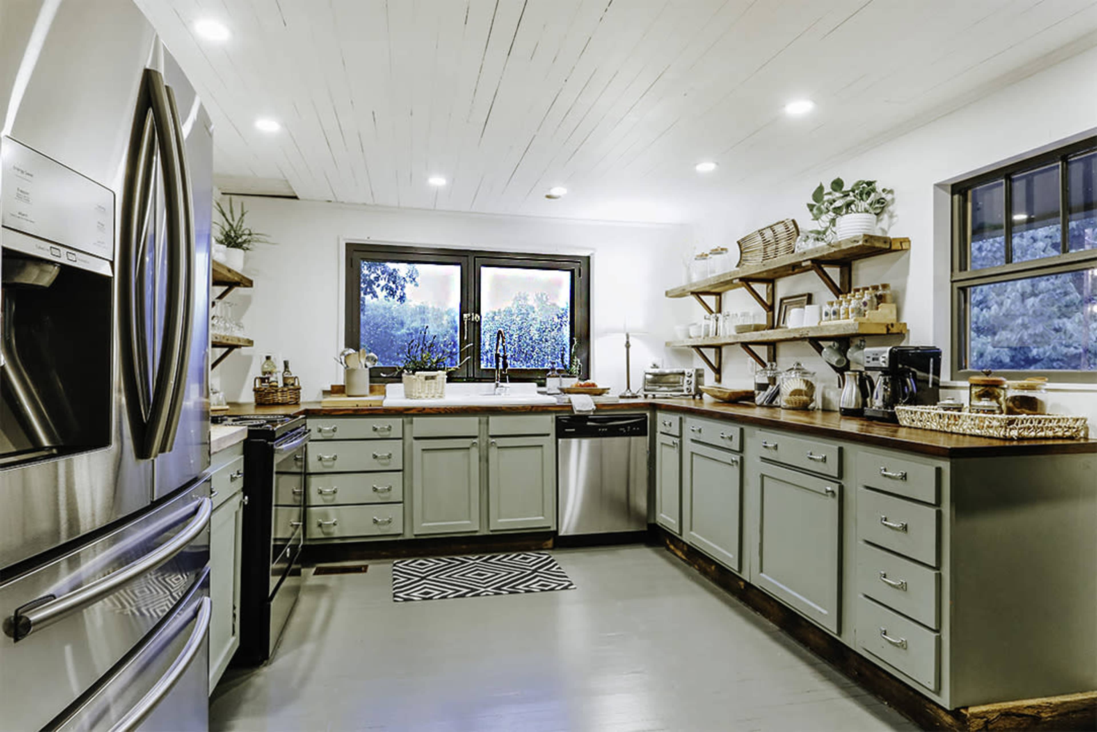 A modern kitchen features gray cabinetry, stainless steel appliances, and open shelving with decorative items, illuminated by overhead lighting.