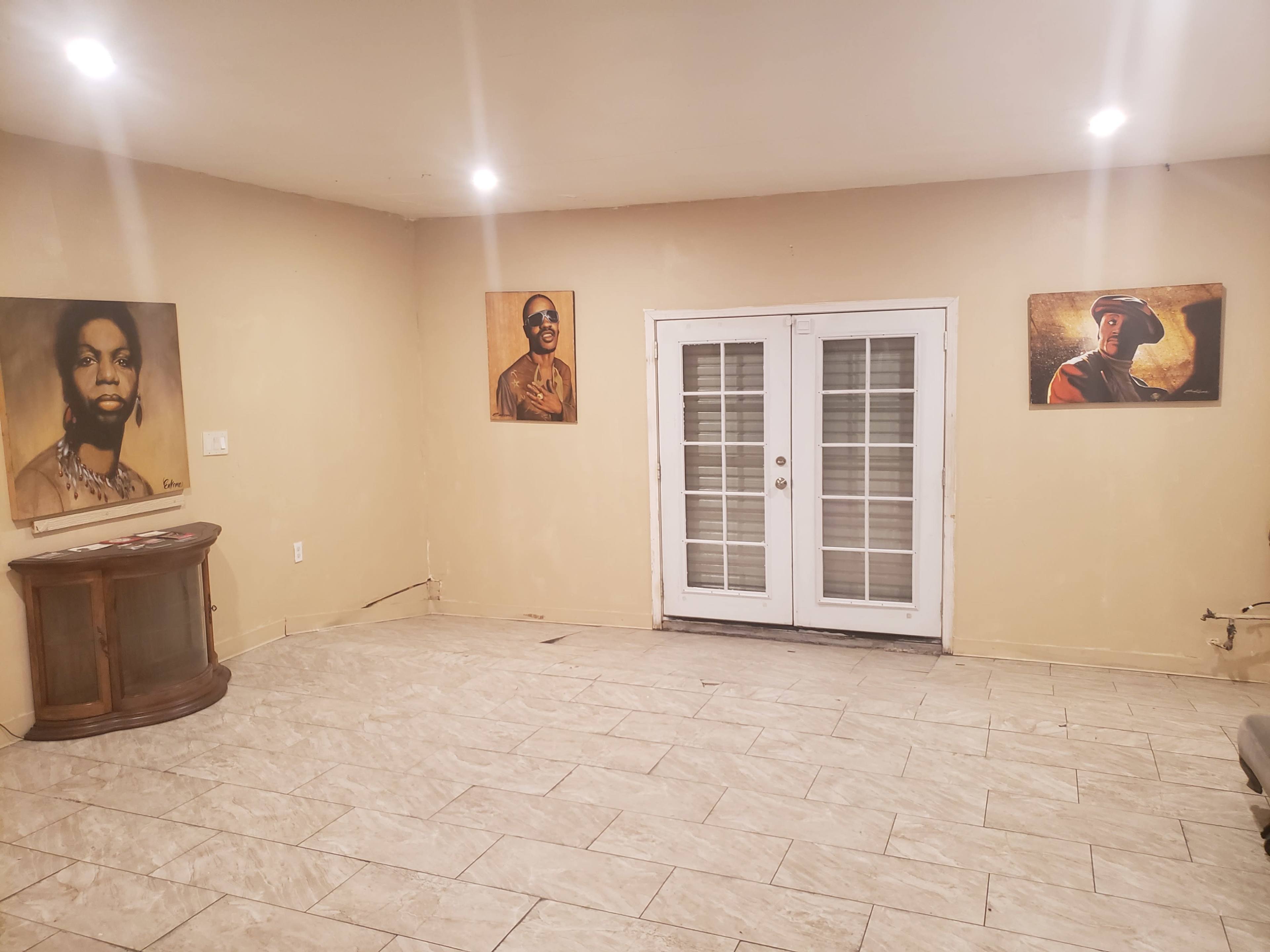 The image shows a sparse room with light-colored walls, tiled flooring, and three framed portraits hanging on the walls, along with a wooden cabinet in one corner.