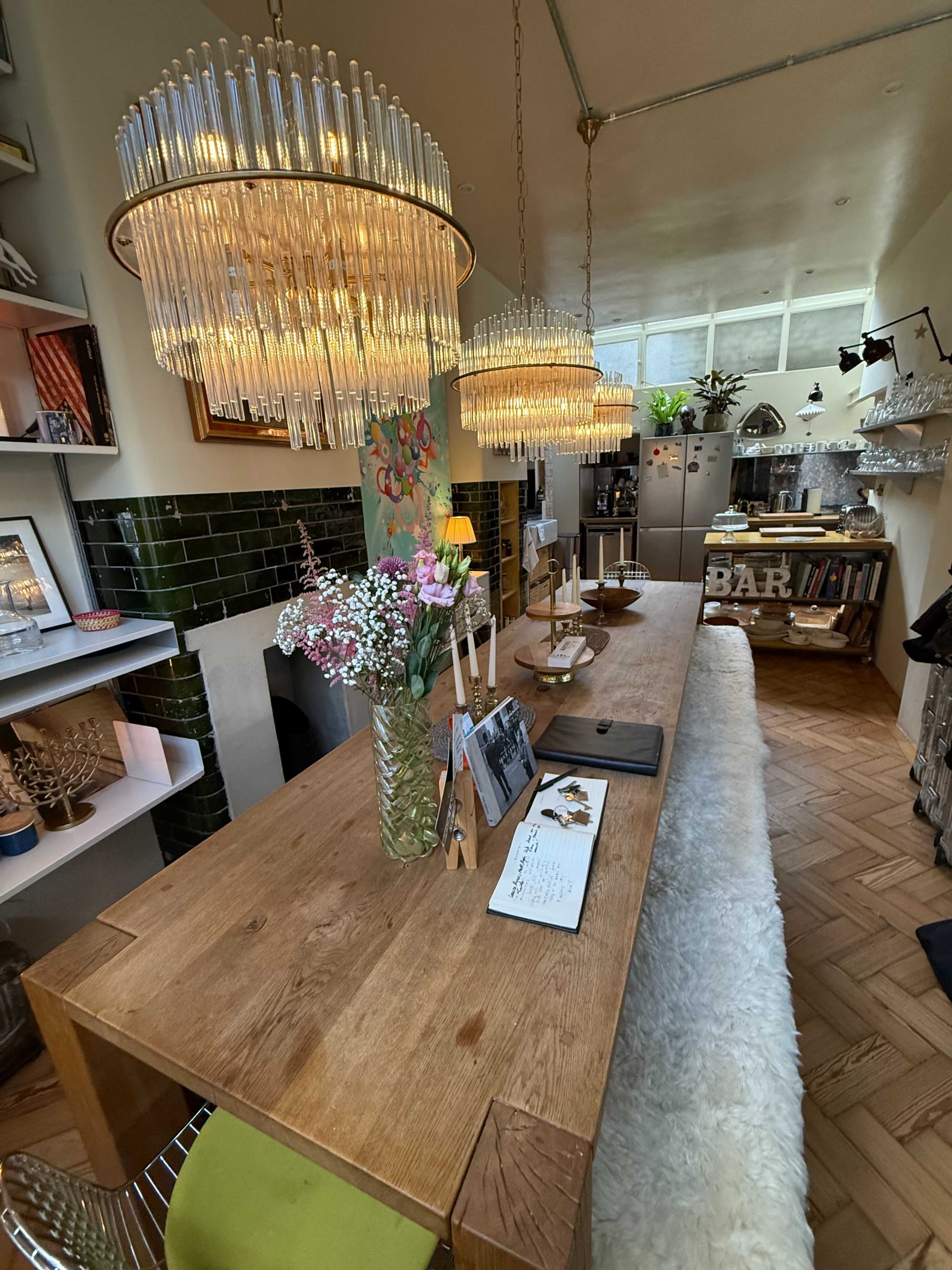 The image shows a cozy cafe interior with a long wooden table, elegant chandeliers, and stylish decor, including flowers and various bookshelves.