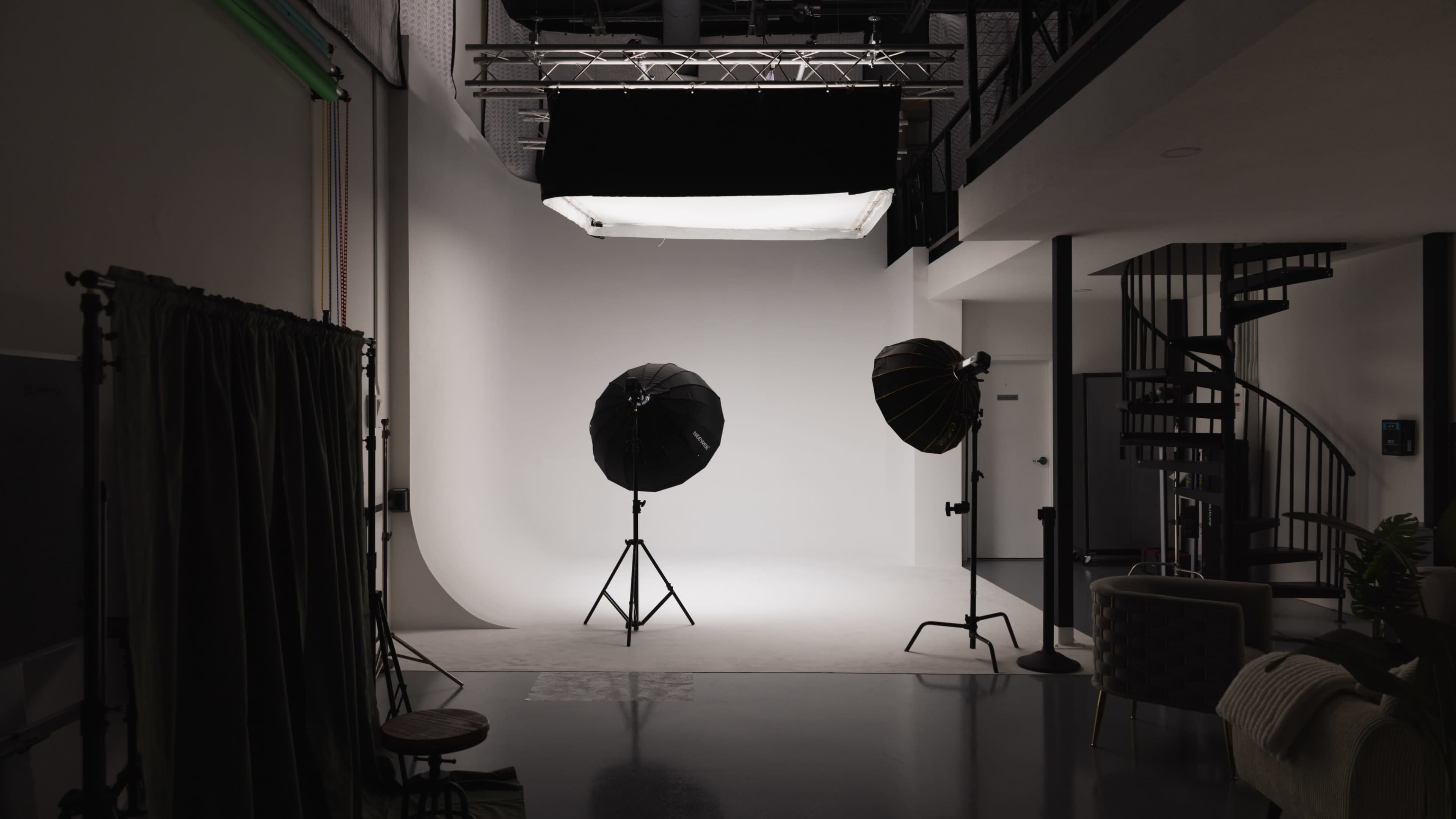 A photography studio features softbox lights on tripods, a seamless backdrop, and a spiral staircase in a minimalistic setting.
