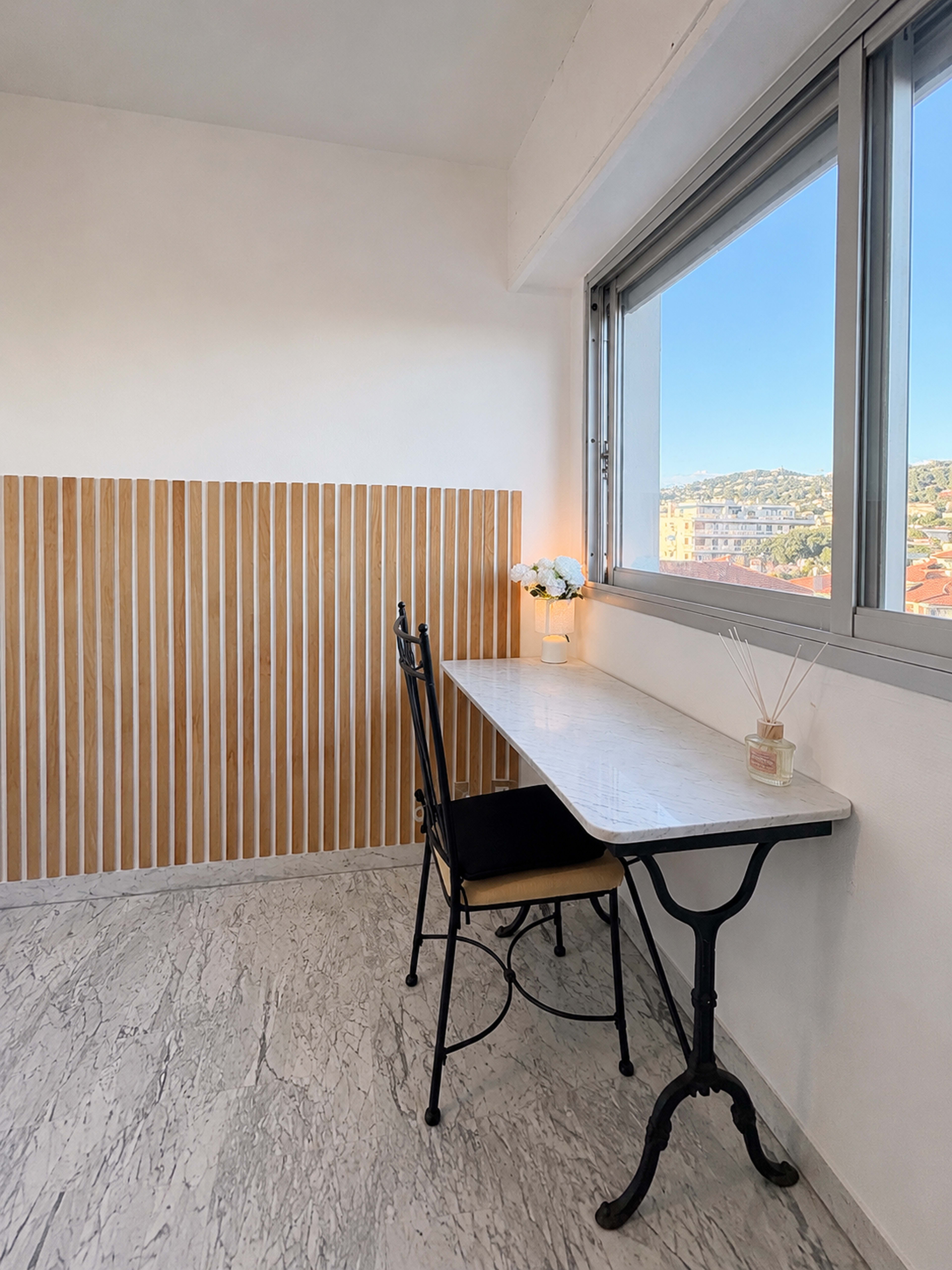 The image shows a minimalist workspace with a marble-top table and a black chair positioned next to a window, featuring a wooden accent wall and a vase of flowers.