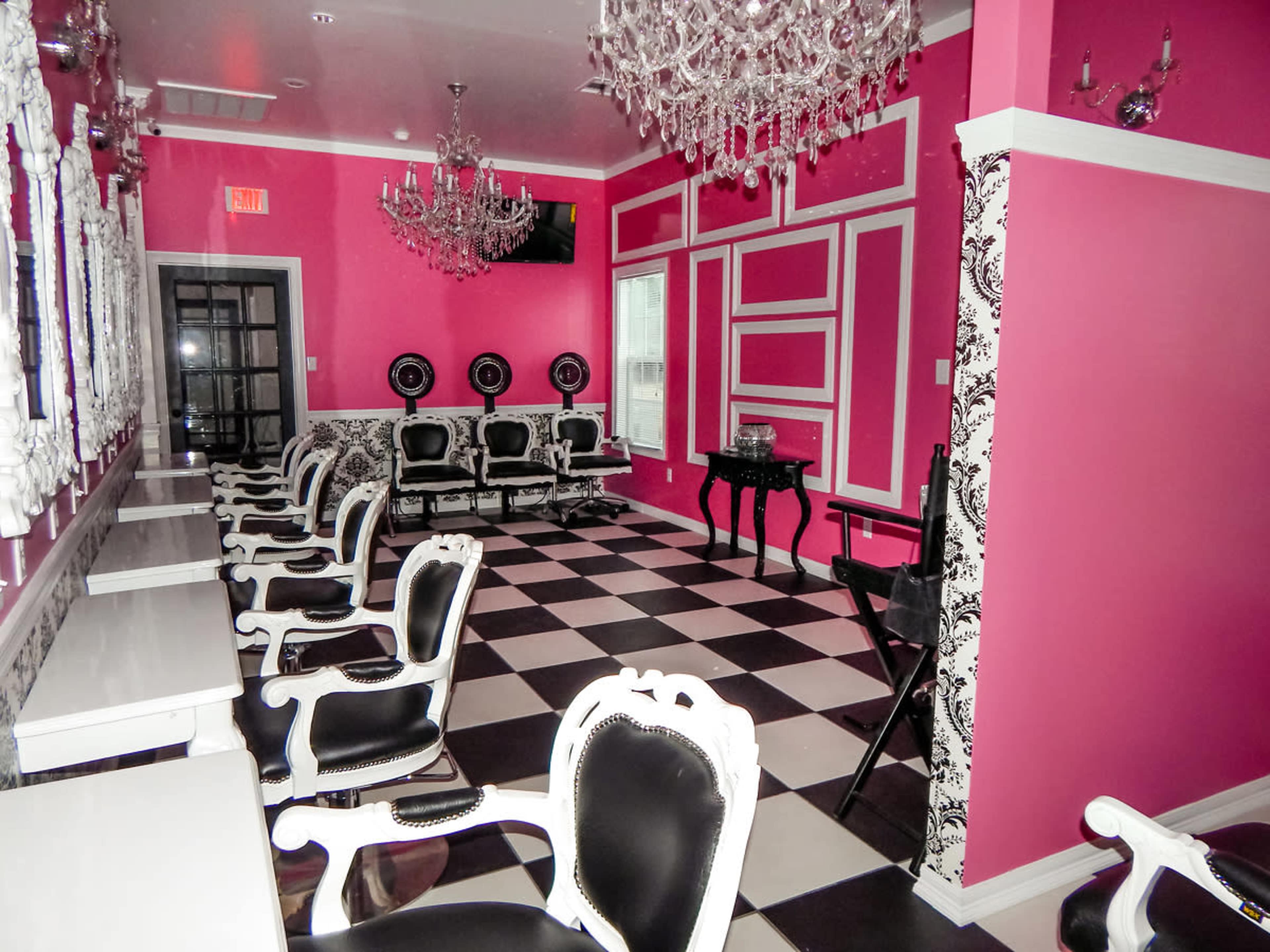 A beauty salon with a pink and black color scheme, featuring chandeliers, black and white checkered flooring, and elegant seating arrangements.