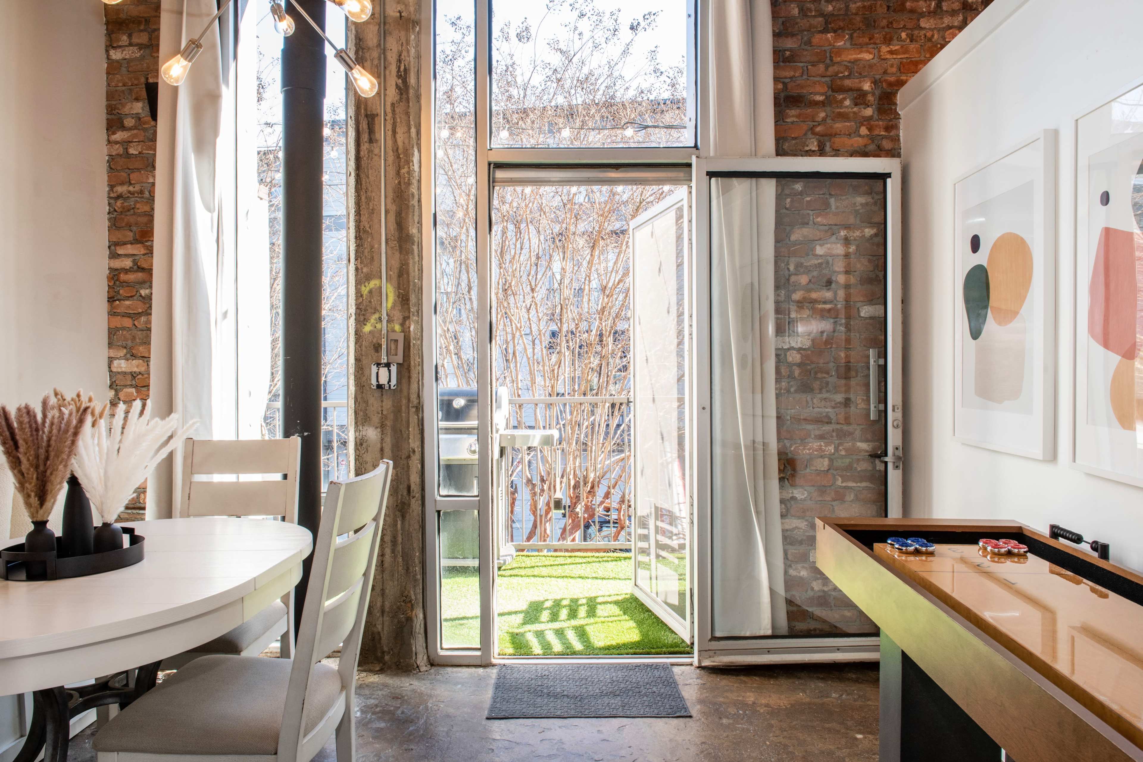 A modern interior space featuring a dining area with a round table, a sliding glass door leading to a balcony, and a small game table along the wall.
