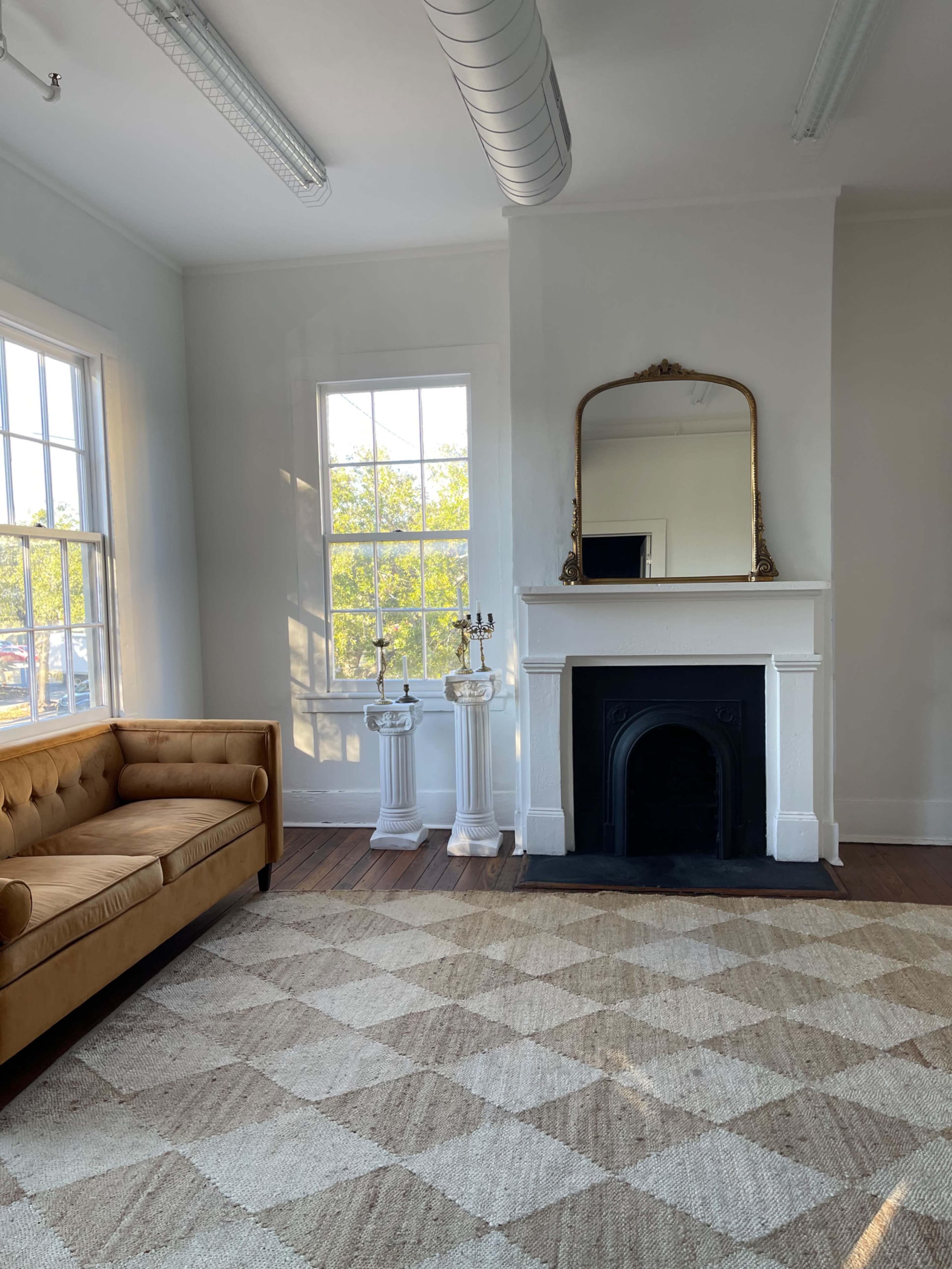 The image shows a bright room featuring a tan sofa, a fireplace with a mirror above it, and two large windows letting in natural light.
