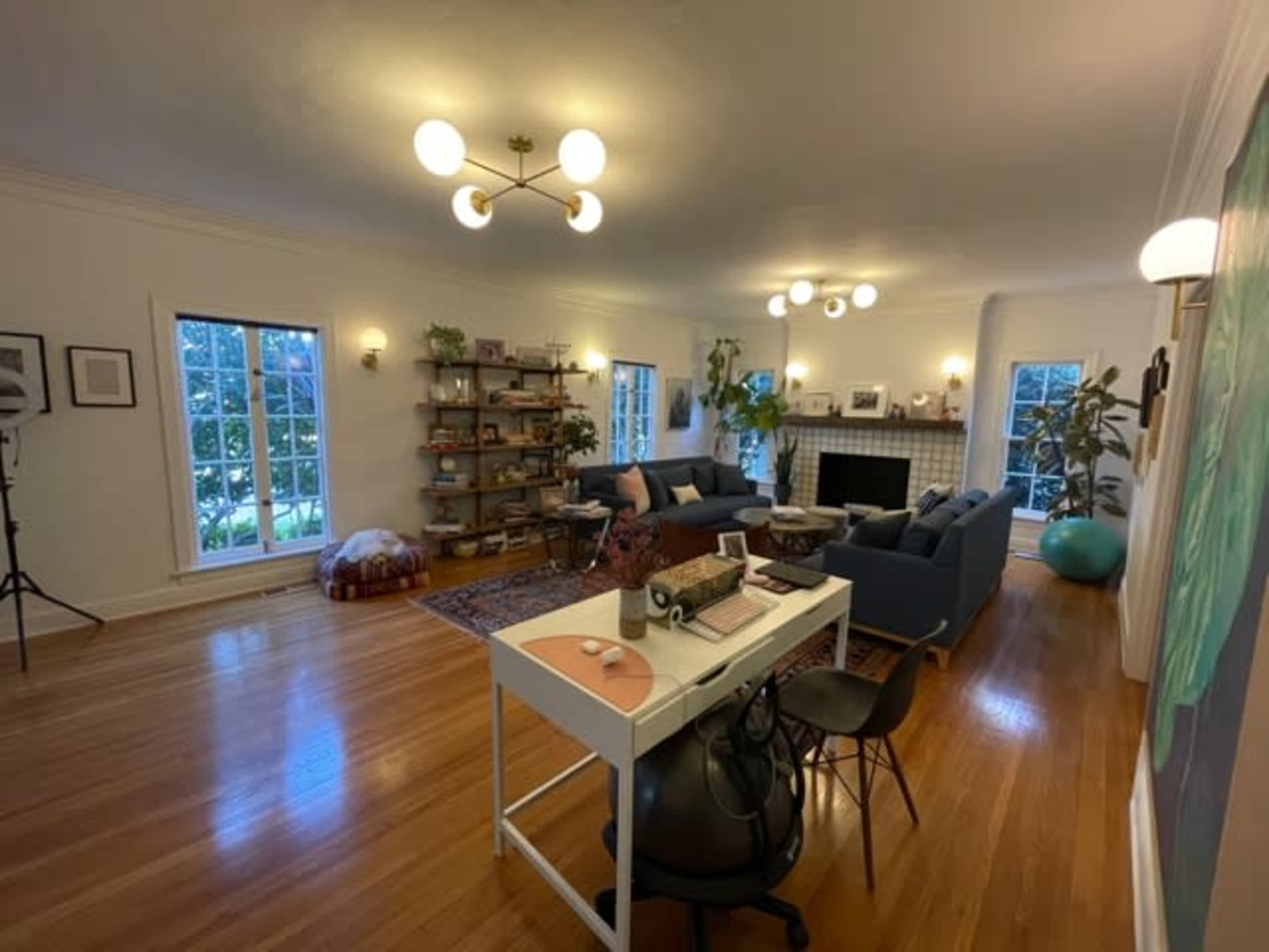 A spacious living room features a seating area with blue sofas, a bookshelf, and a desk, alongside large windows allowing natural light to fill the space.