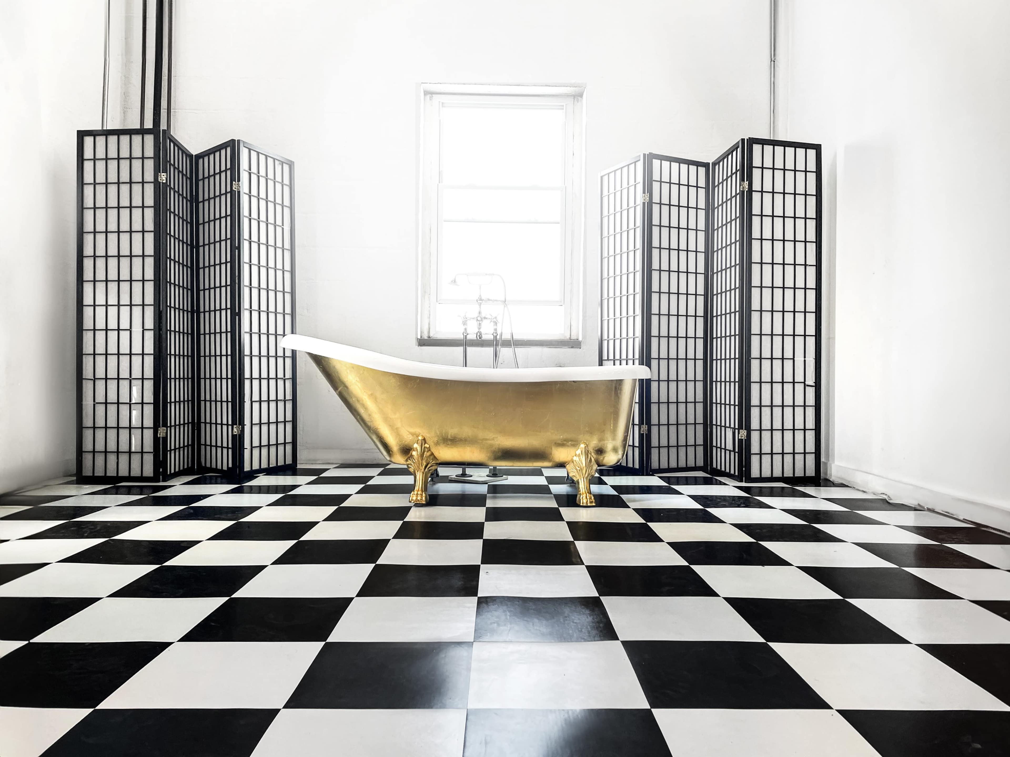 A gold and white freestanding bathtub sits on a black-and-white checkered floor, surrounded by three black folding screens in a bright, minimalistic room.