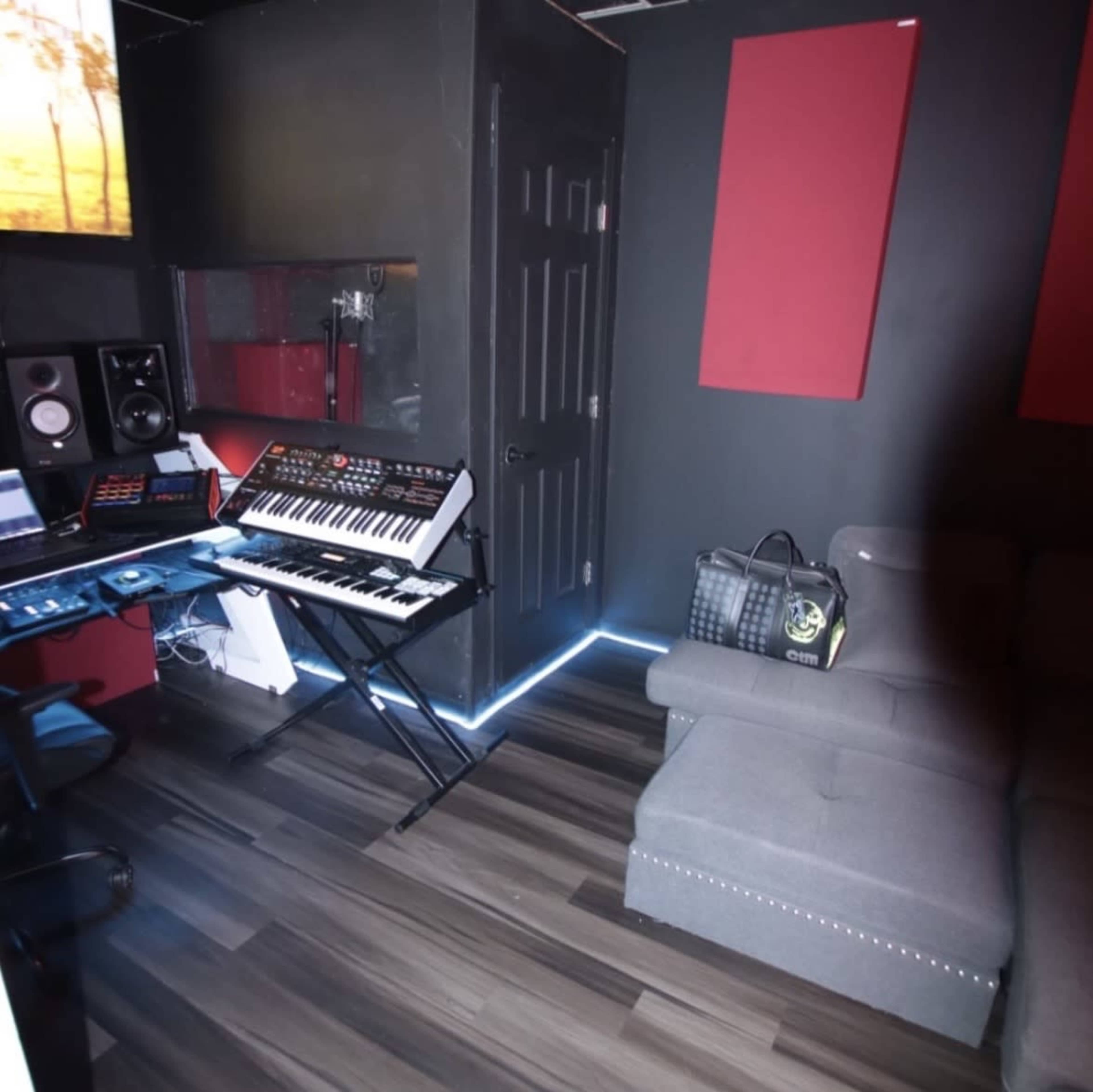 The image shows a music studio with a keyboard, audio equipment, and a gray couch in a dimly lit room featuring red soundproof panels and wooden flooring.