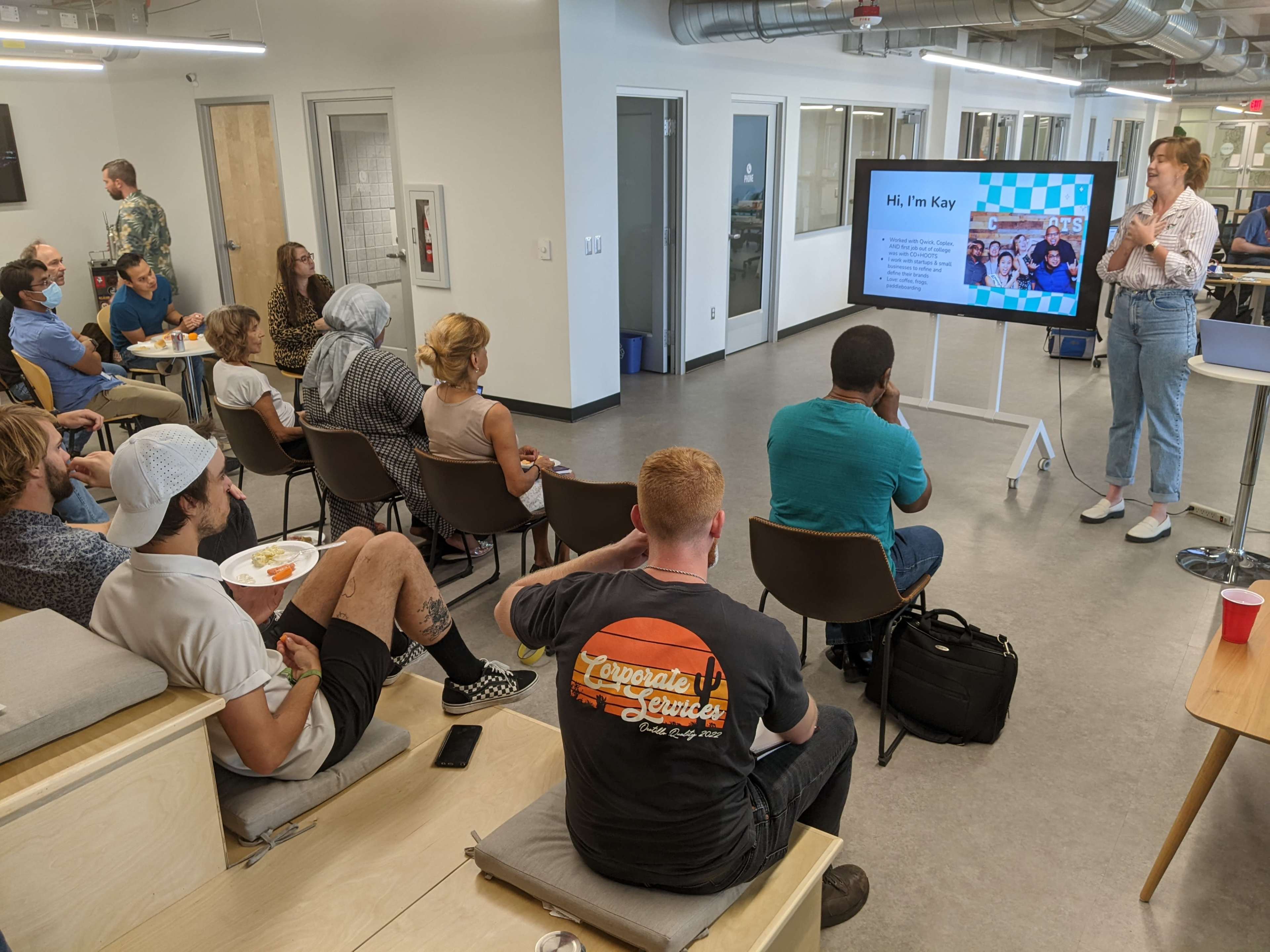 A speaker stands in front of a seated audience in a modern office space, presenting slide content on a large screen.