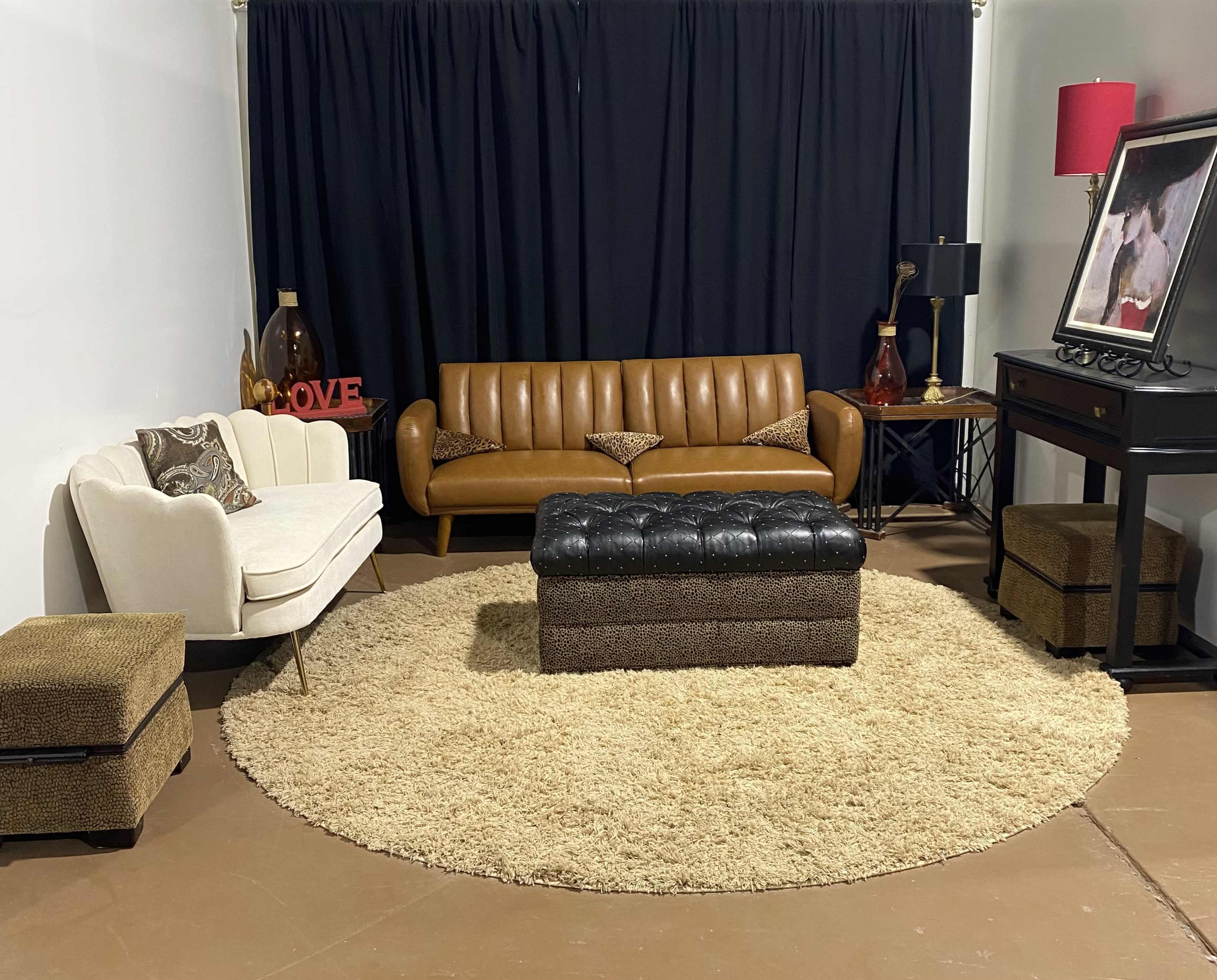 The image shows a living room setup featuring a brown leather sofa, a white armchair, a round area rug, a black ottoman, and a console table with a lamp and decorative items against a black curtain backdrop.