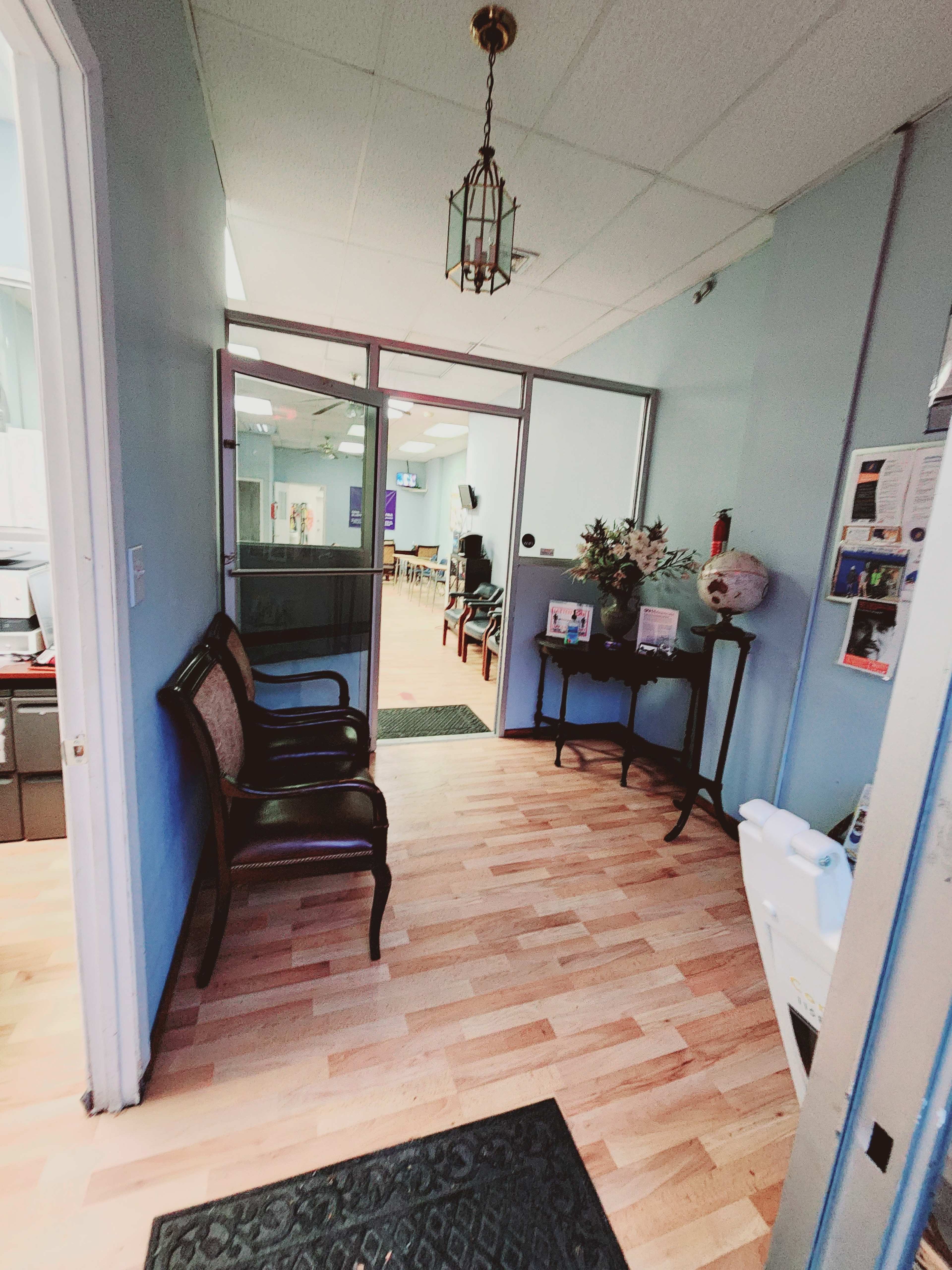 A hallway leading to a glass door, with wooden flooring, a small table with decor, and two chairs against the wall.