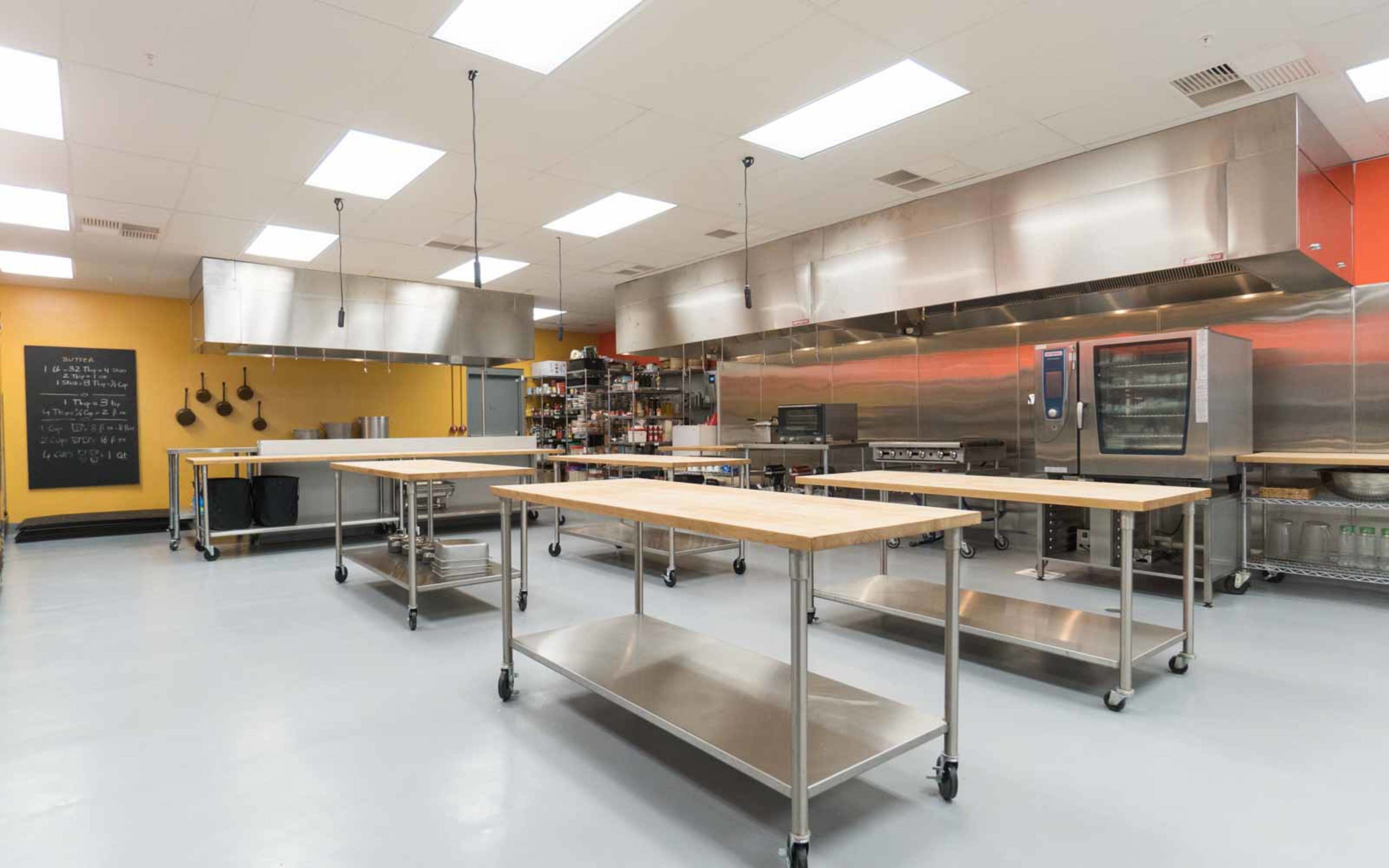 A spacious commercial kitchen with stainless steel equipment, multiple workstations with wooden countertops, and organized shelves of supplies.