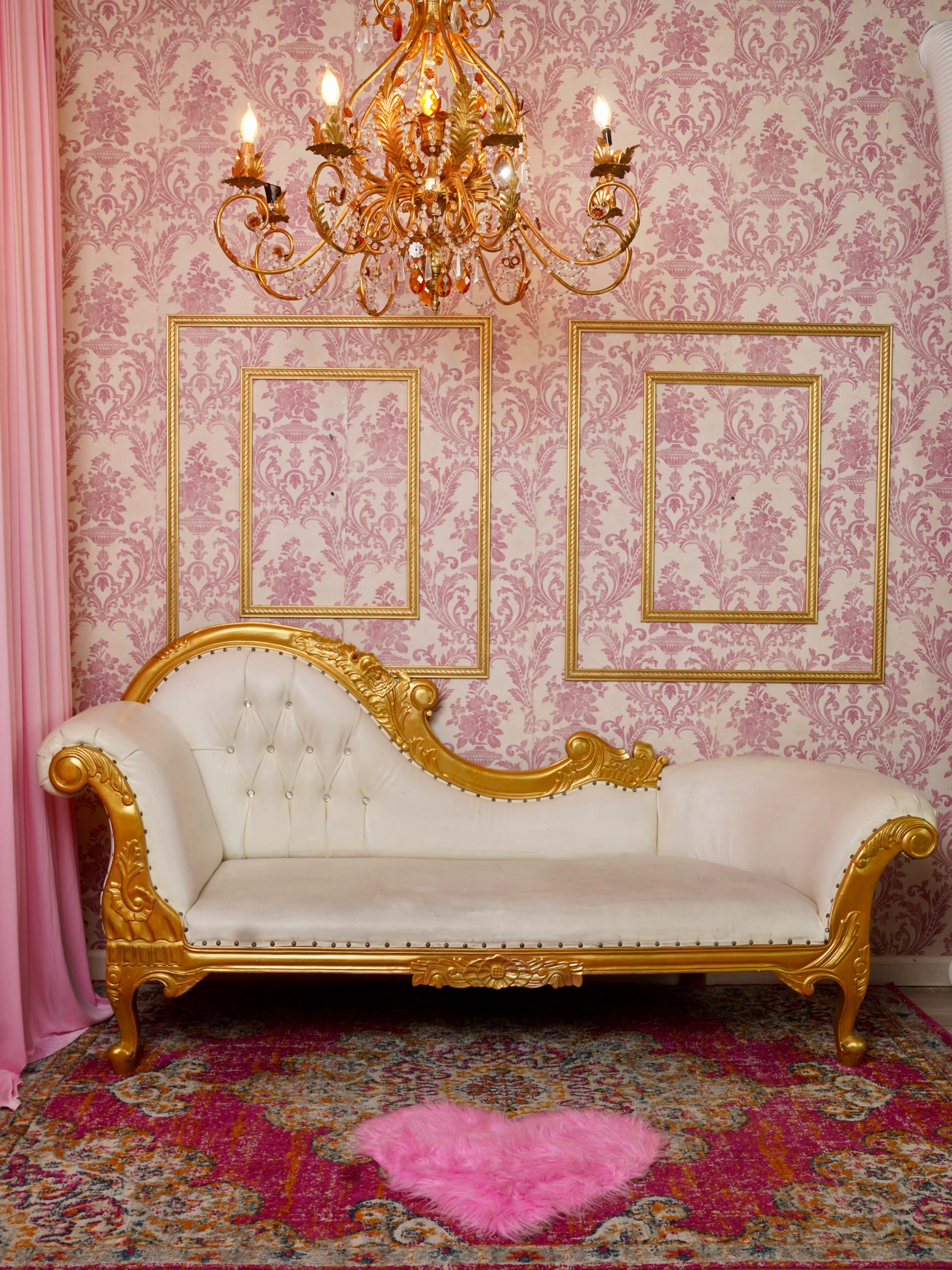 The image features an ornate gold and white chaise longue placed against a pink floral-patterned wall, beneath a luxurious chandelier, with a heart-shaped pink rug on the floor.
