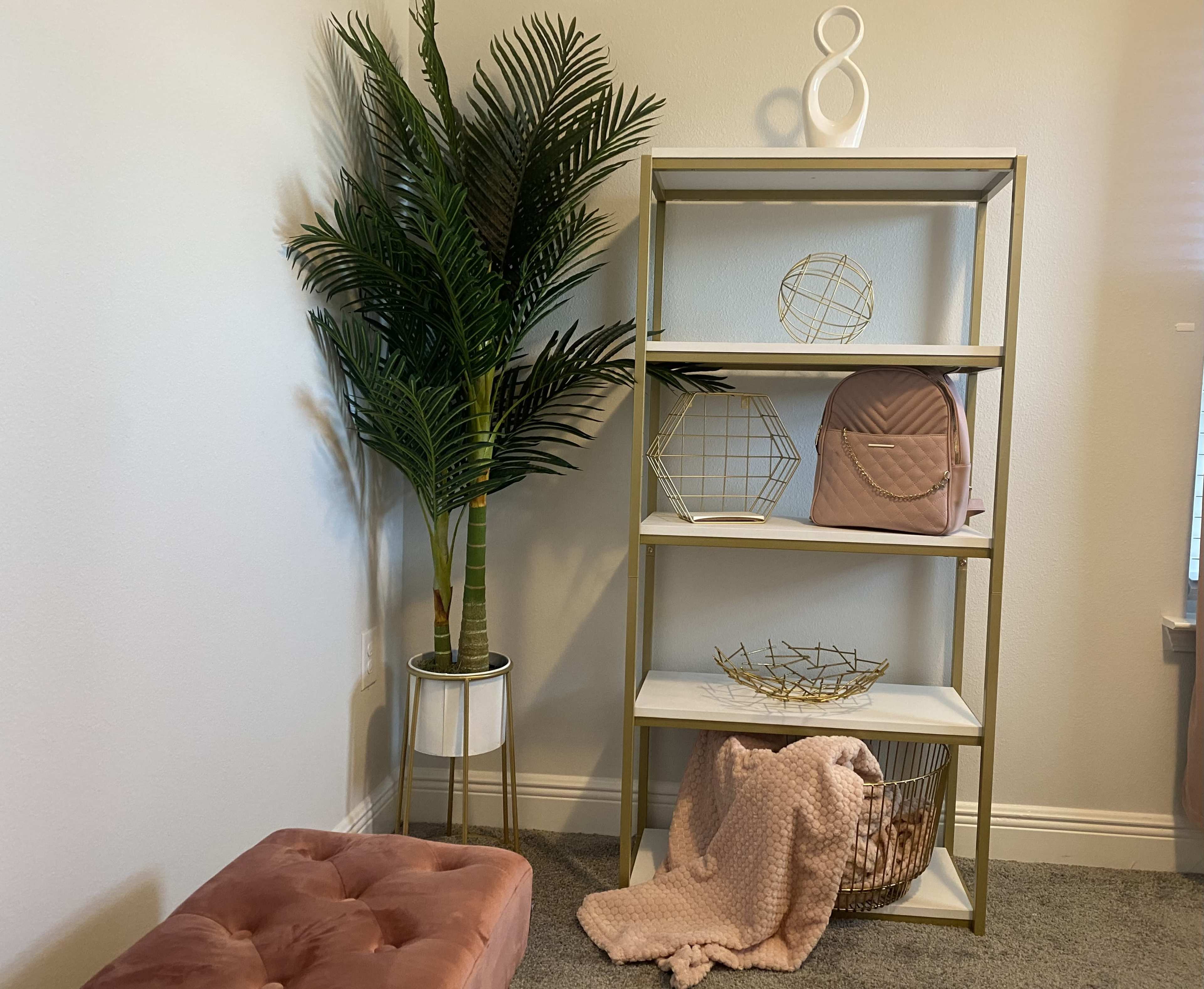 A light-colored room with a metal bookshelf featuring decorative items, a tall potted plant, a pink backpack, and a plush pink ottoman.