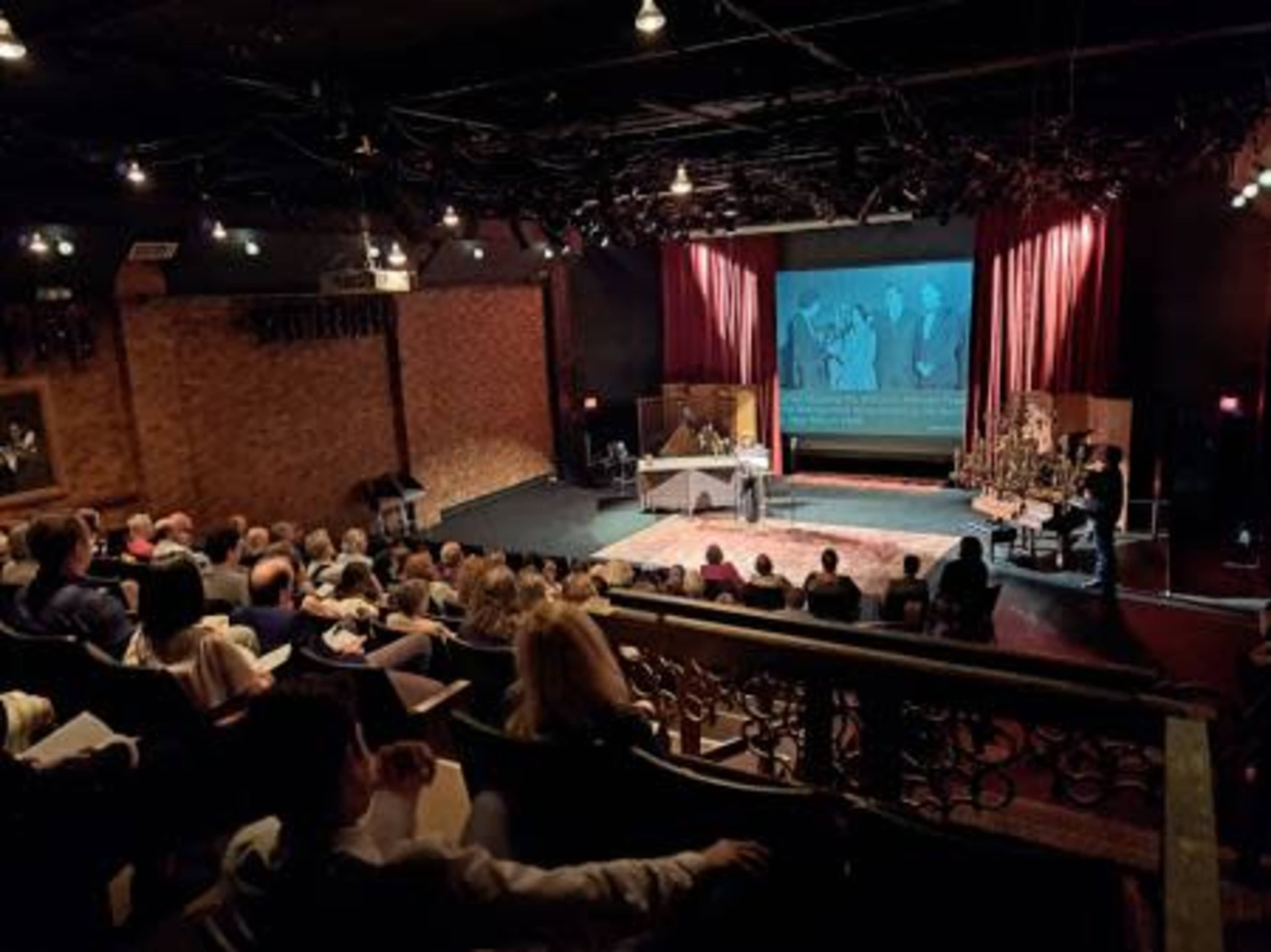 A theater audience watches a presentation on stage with a large screen displaying black-and-white images.