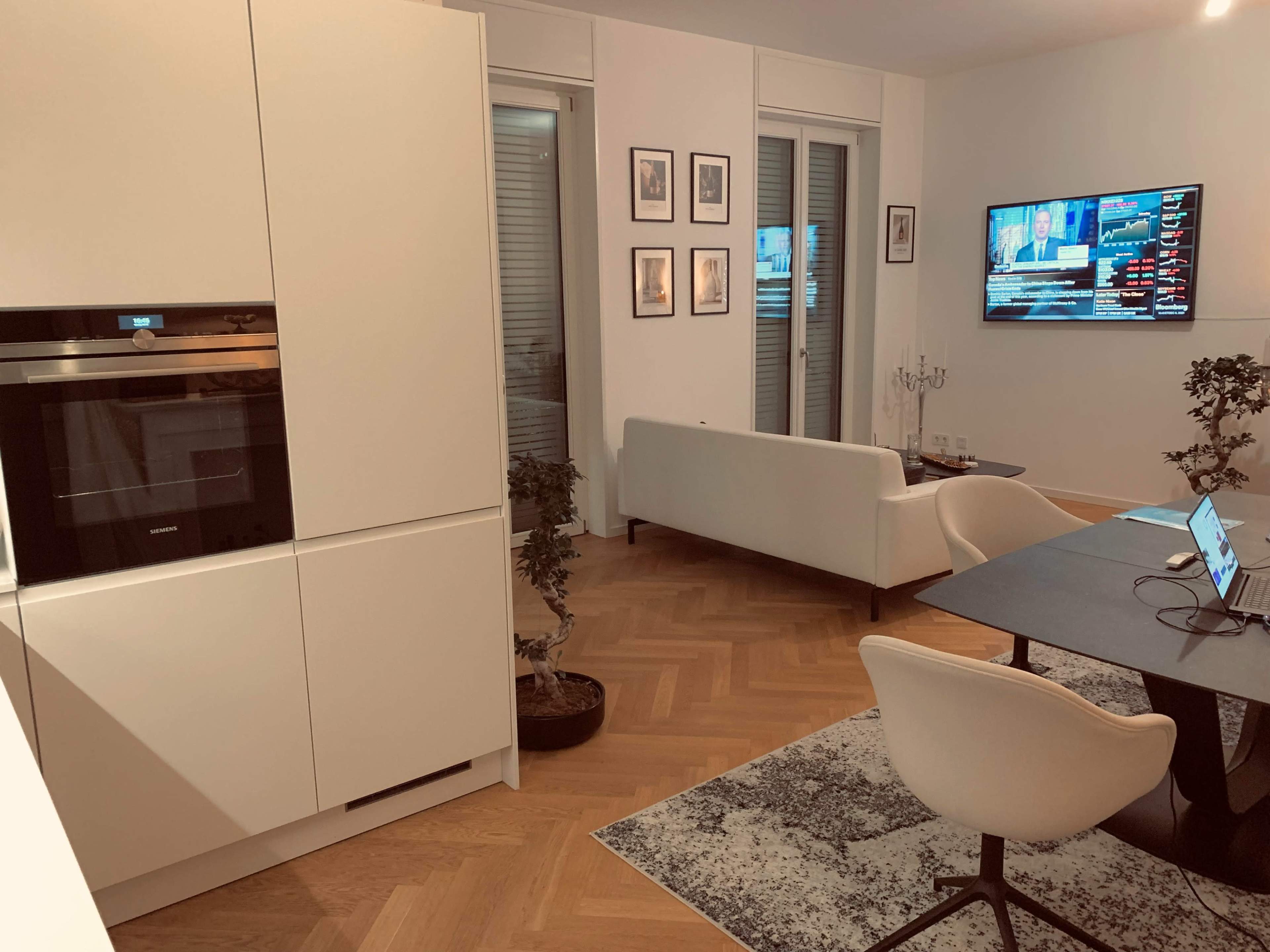 A modern living space features a white sofa, a dining table with chairs, and an oven in a sleek kitchen, with a television displaying news in the background.