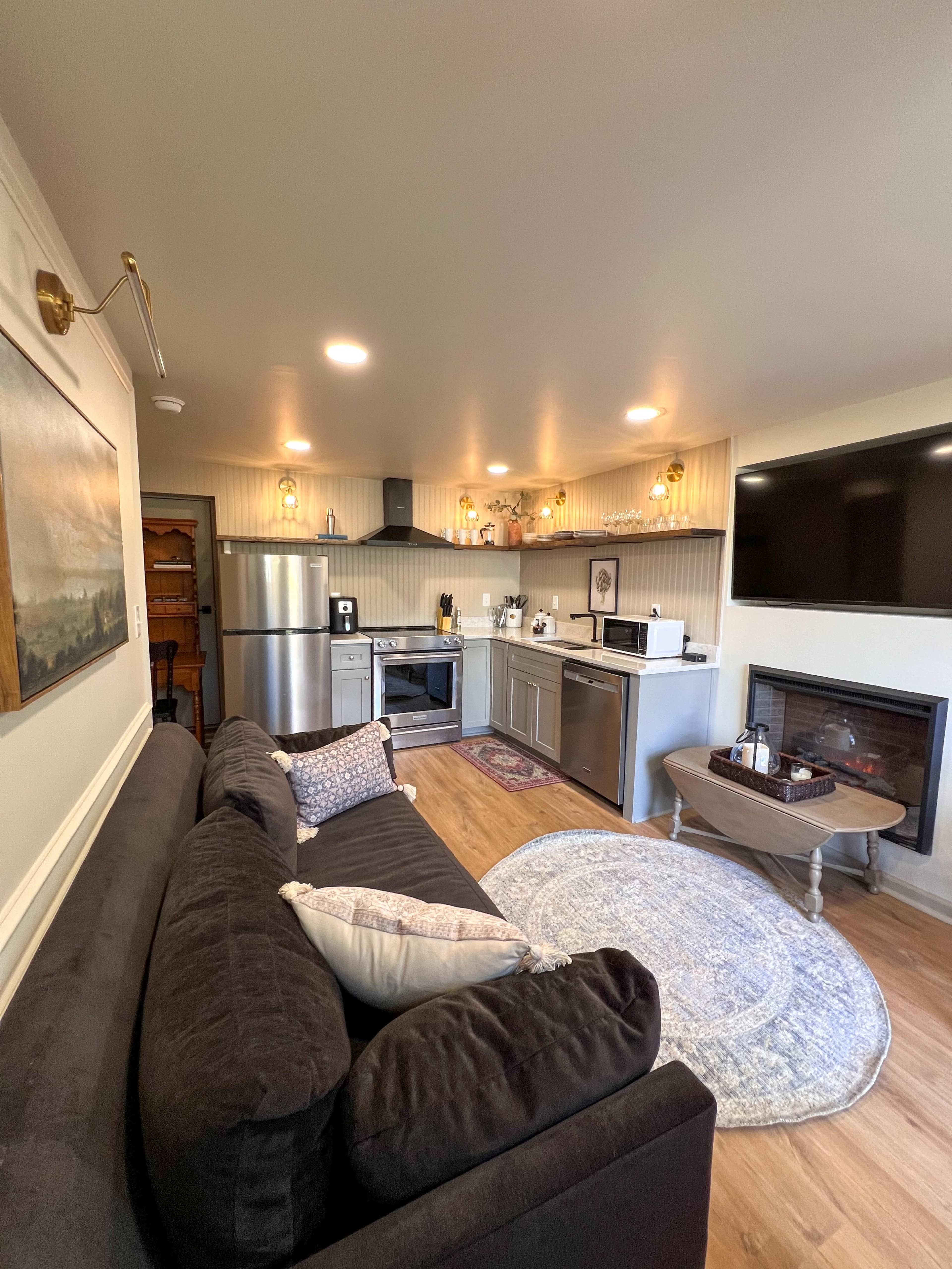A modern kitchen and living area with stainless steel appliances, a fireplace, and a couch facing a wall-mounted television.