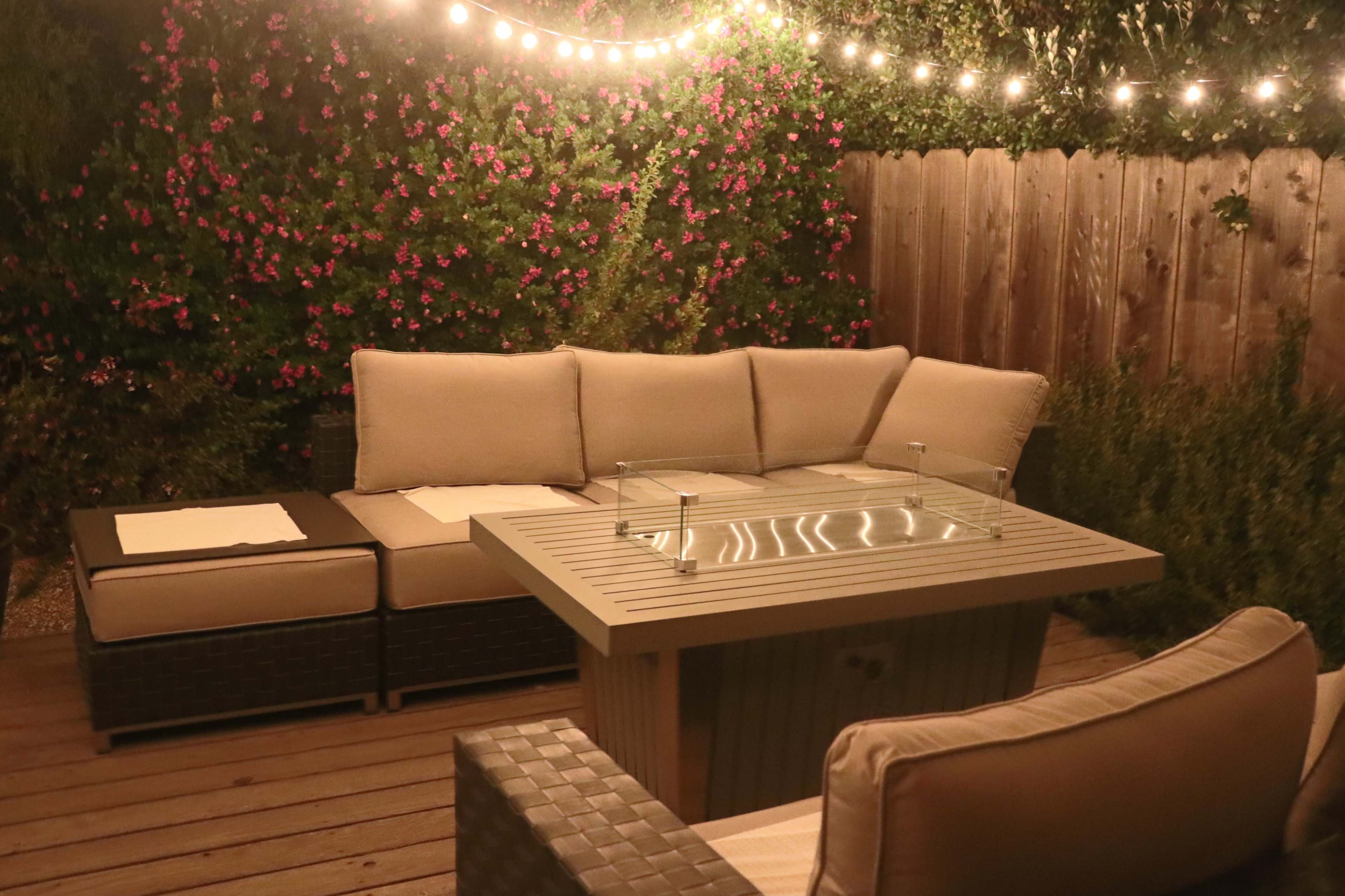 A cozy outdoor seating area features a sectional sofa and a fire pit table, illuminated by string lights against a backdrop of flowering plants.