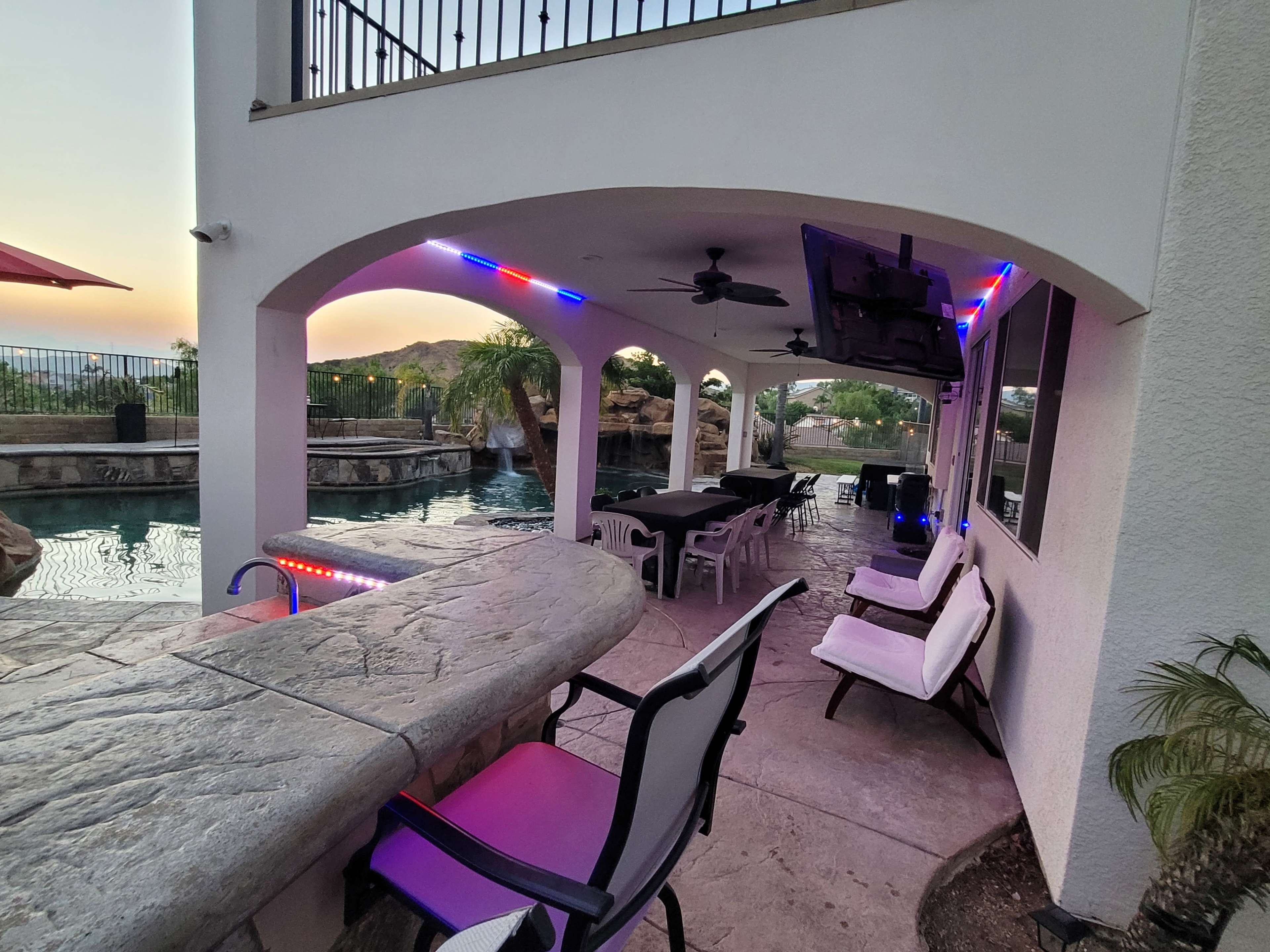 A patio area near a pool, featuring a bar with seating, a television mounted on the wall, and colorful lights along the ceiling.