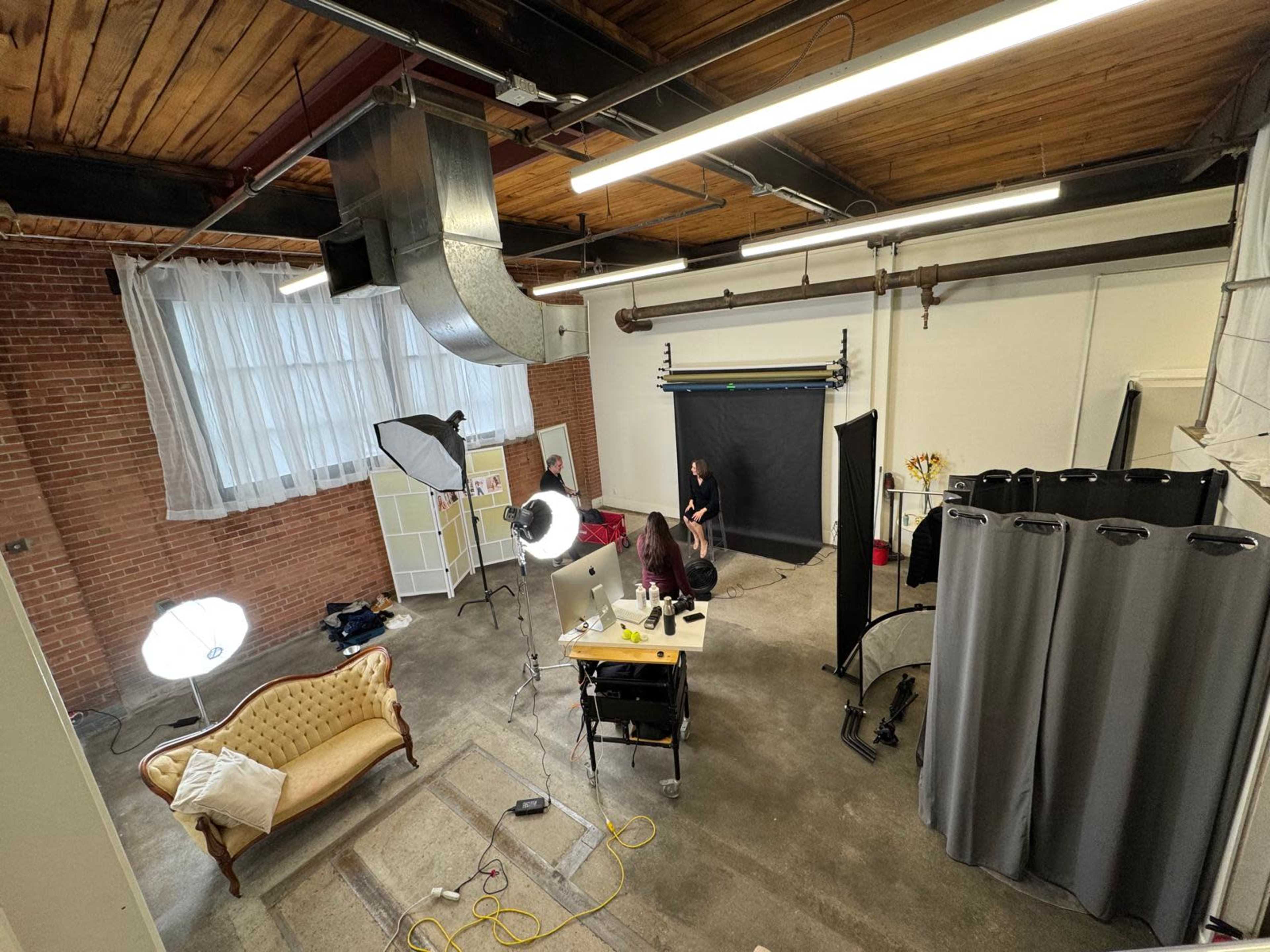 The image shows a spacious studio with a vintage sofa, lighting equipment, a backdrop for photography, and a work desk with a computer.