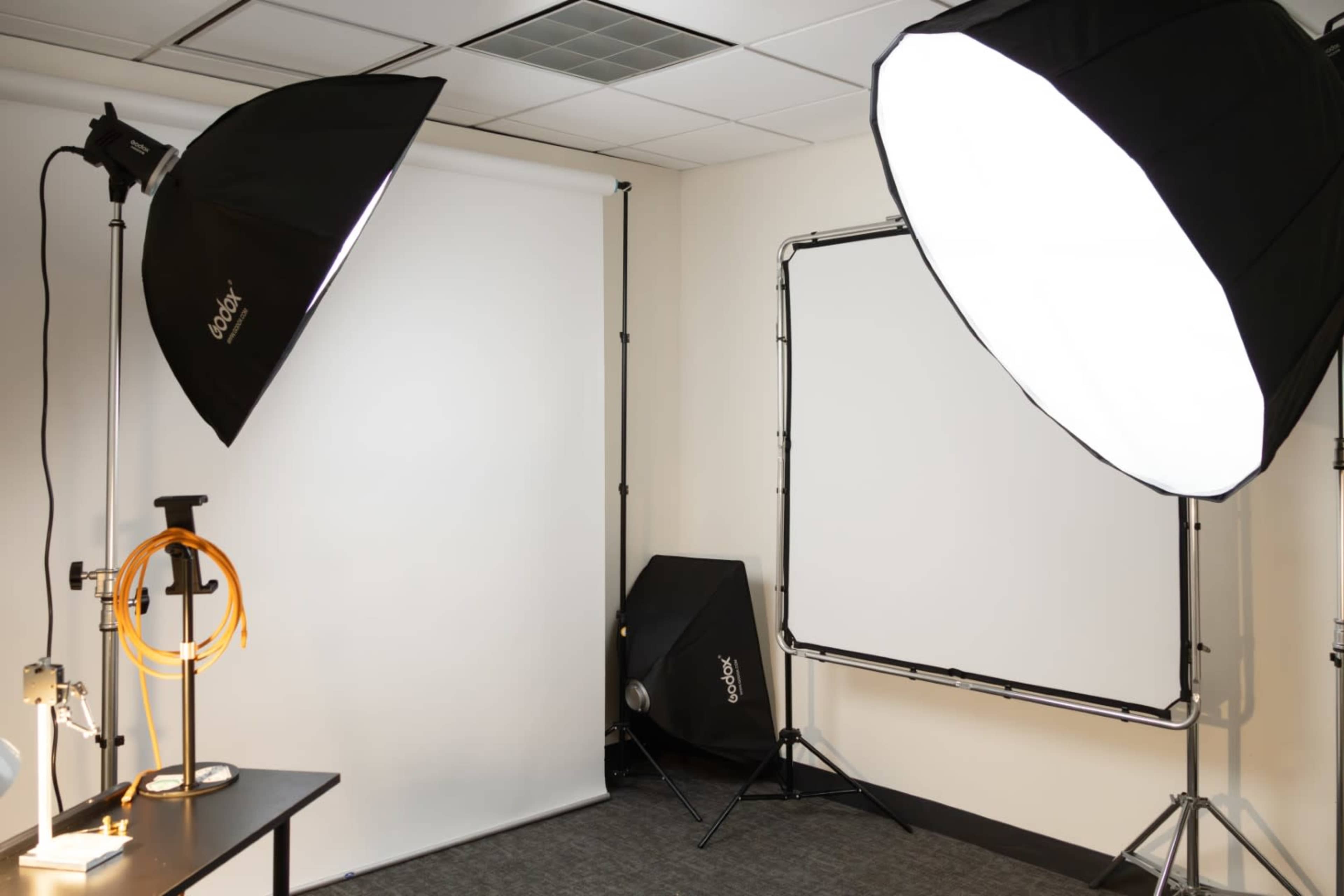 A photography studio setup featuring two softbox lights, backdrop equipment, and a table with lighting accessories.