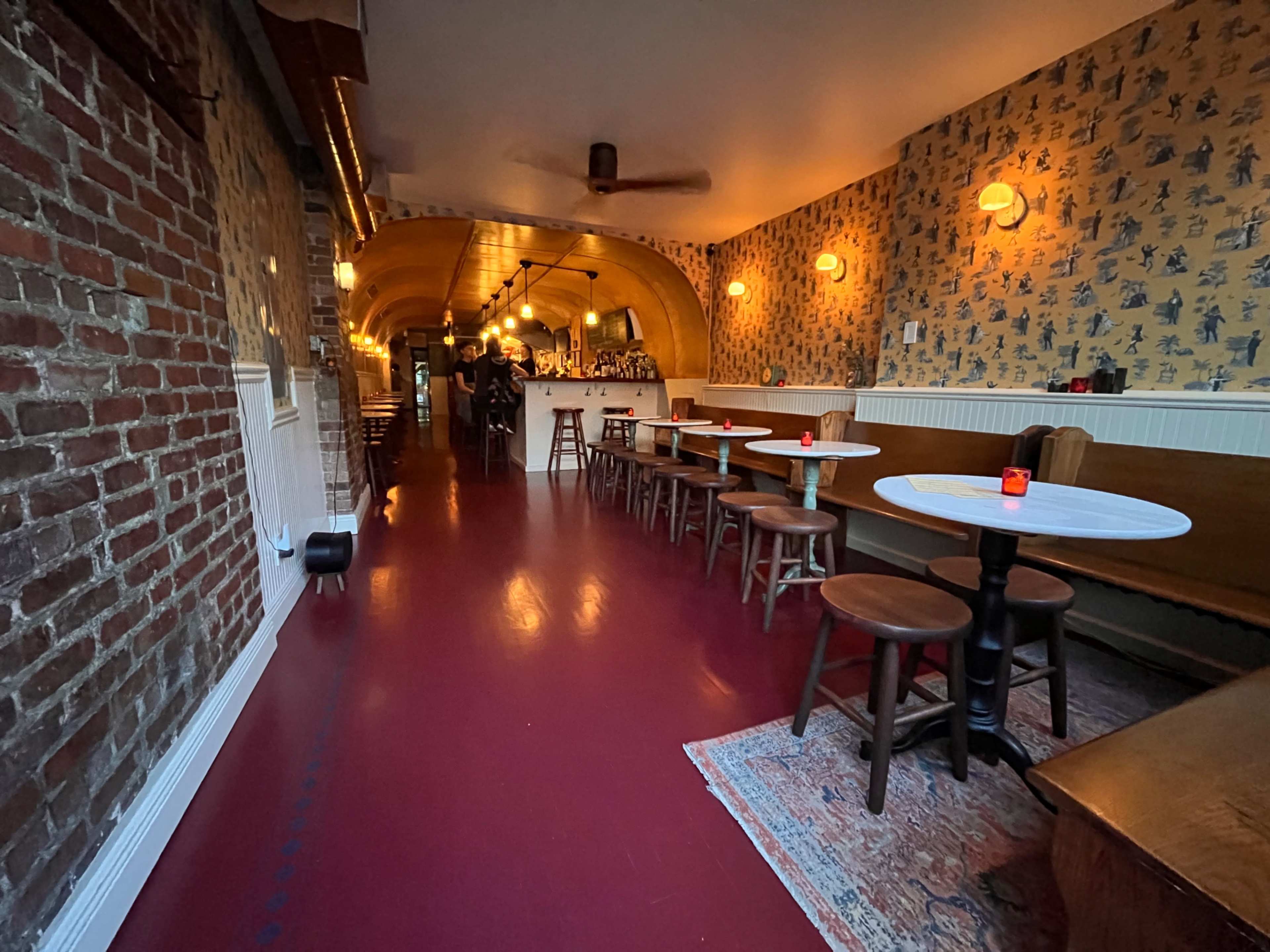 The image shows an interior of a restaurant with brick and patterned walls, wooden tables, and chairs along a narrow, red floor hallway leading to a bar area.