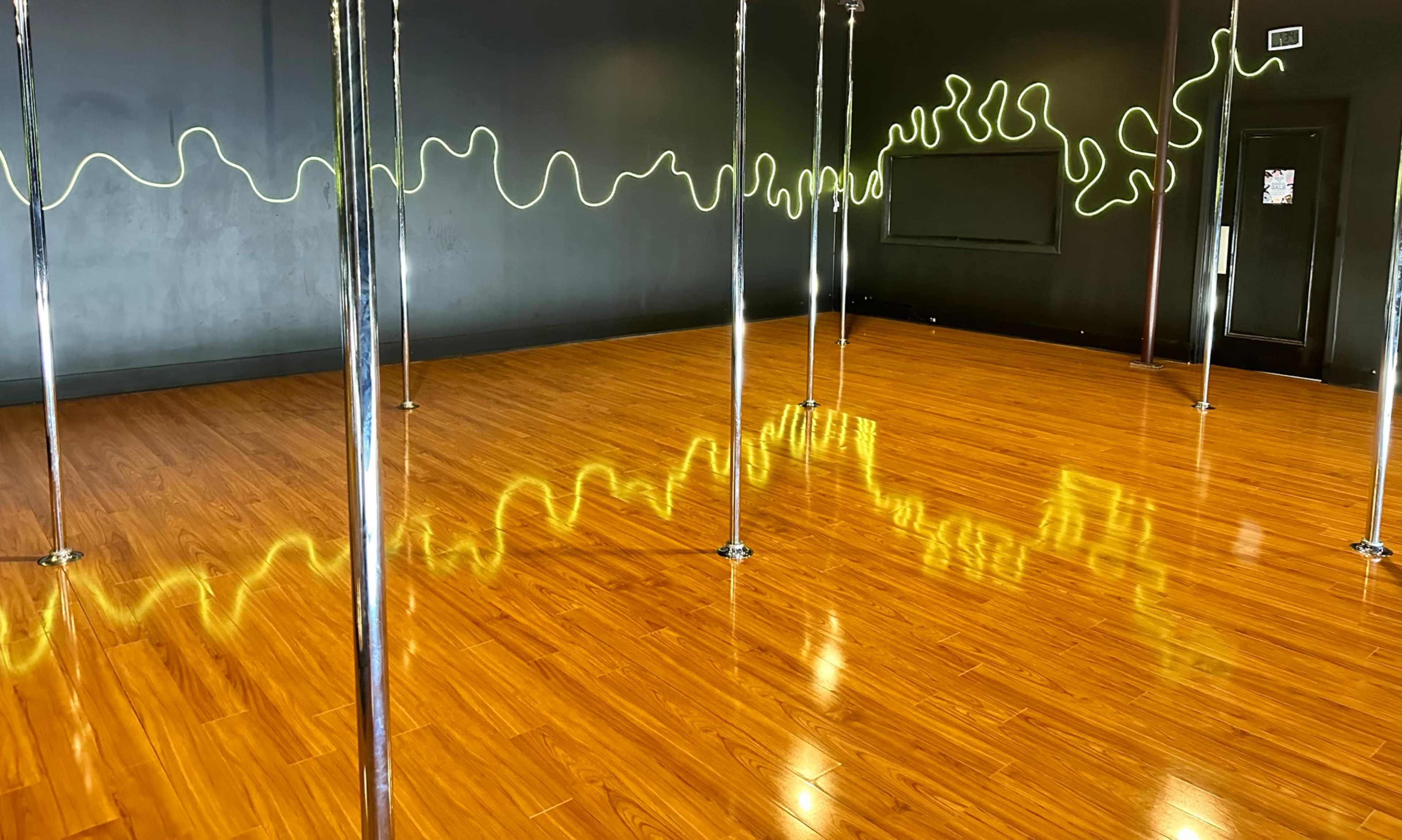 A spacious dance studio with polished wooden floors, illuminated by a neon yellow line on the black wall and several metallic poles placed throughout the room.