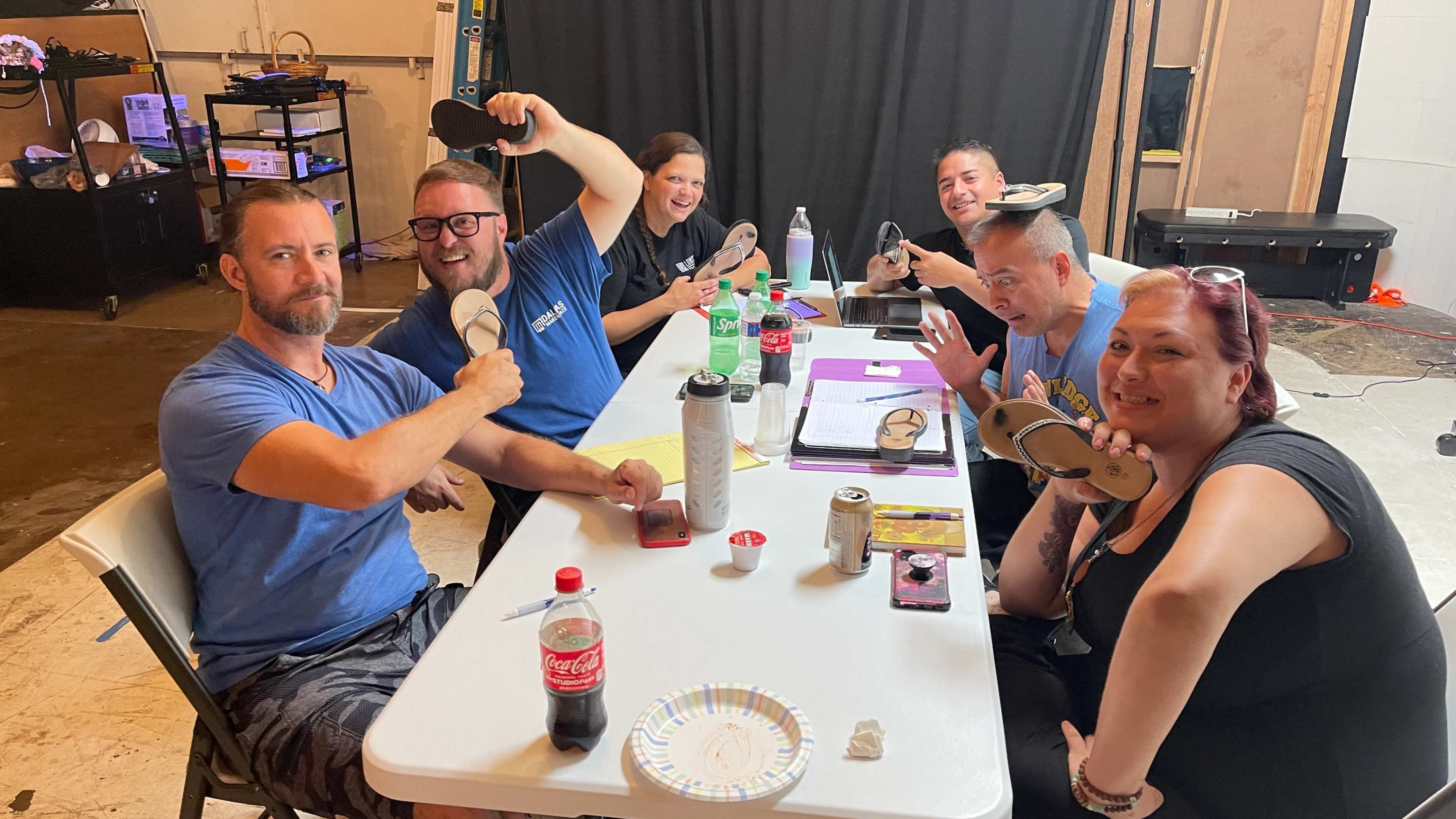 A group of six people sit around a table holding up various flip-flops and drinks while smiling at the camera.