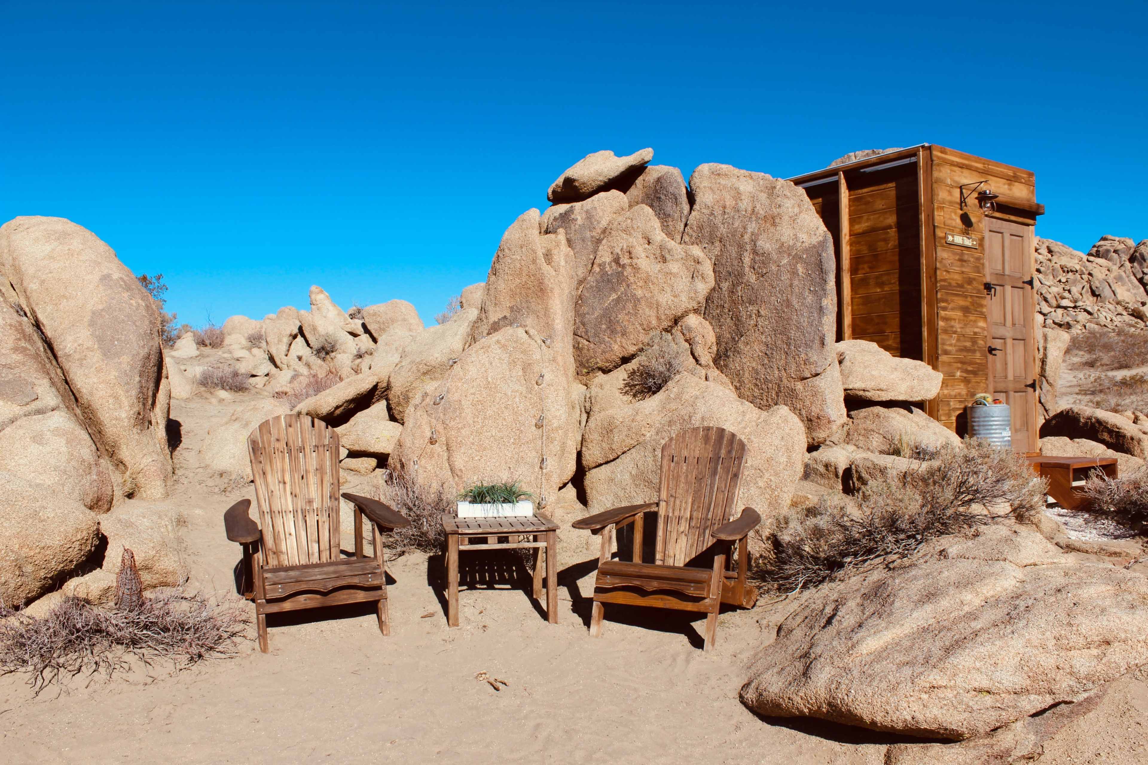 Two wooden chairs and a small table set between large rock formations in a desert landscape, with a wooden structure visible in the background.