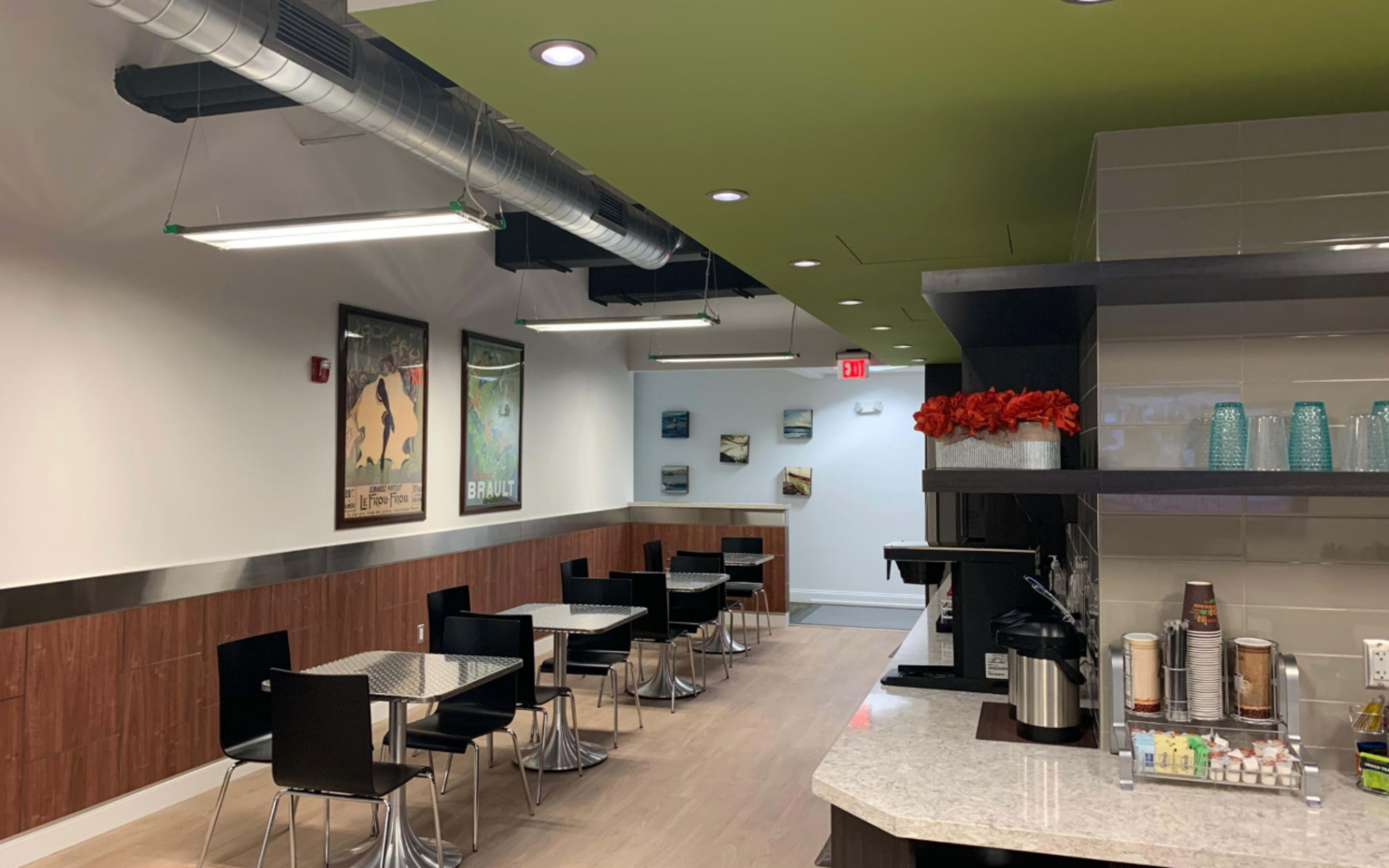 The image shows a modern, well-lit café interior with tables and chairs arranged along a wall and a coffee station at one end.
