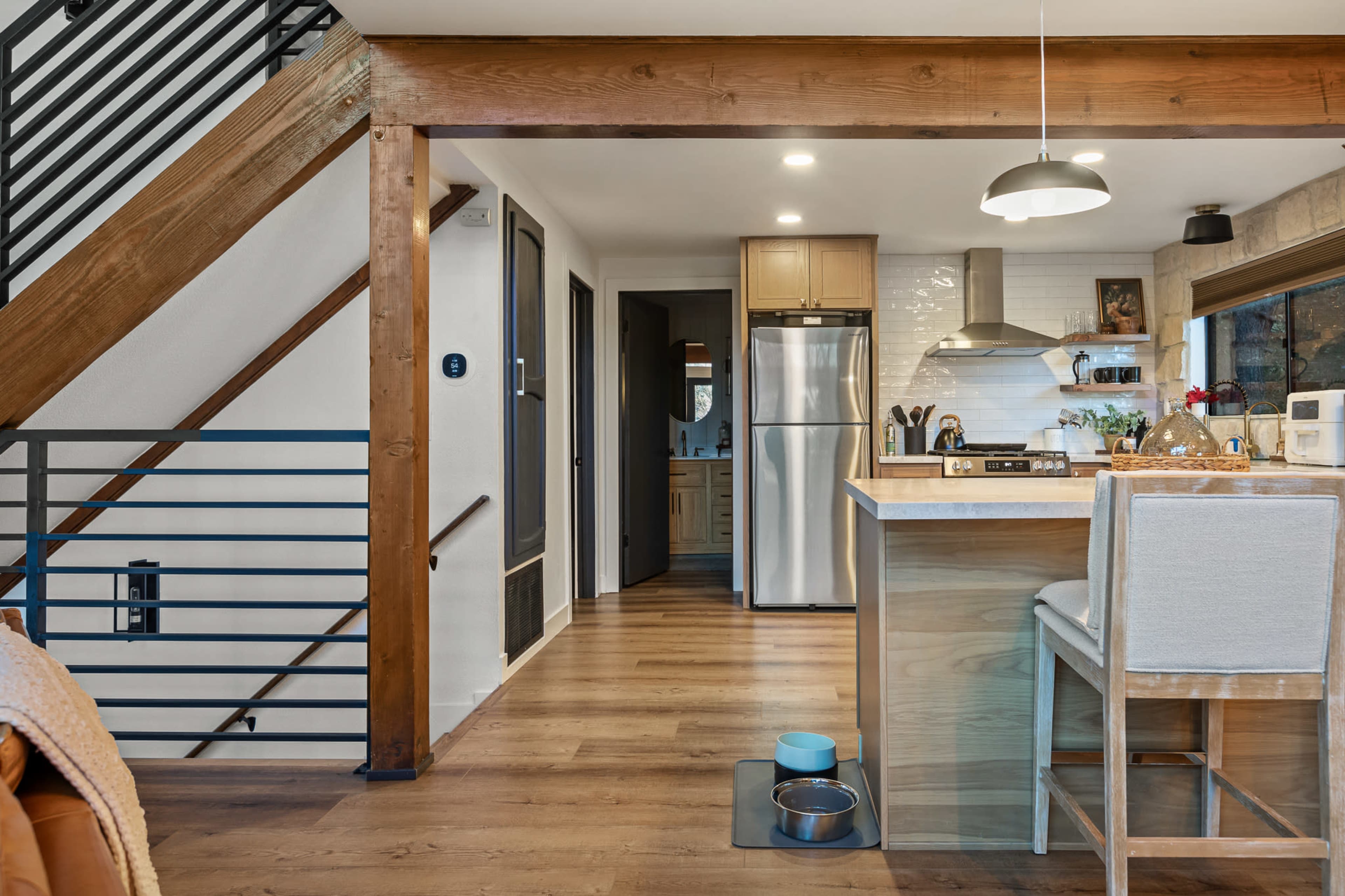 The image shows a modern kitchen area with a staircase in the background, featuring wooden beams, stainless steel appliances, and a small dining setup.