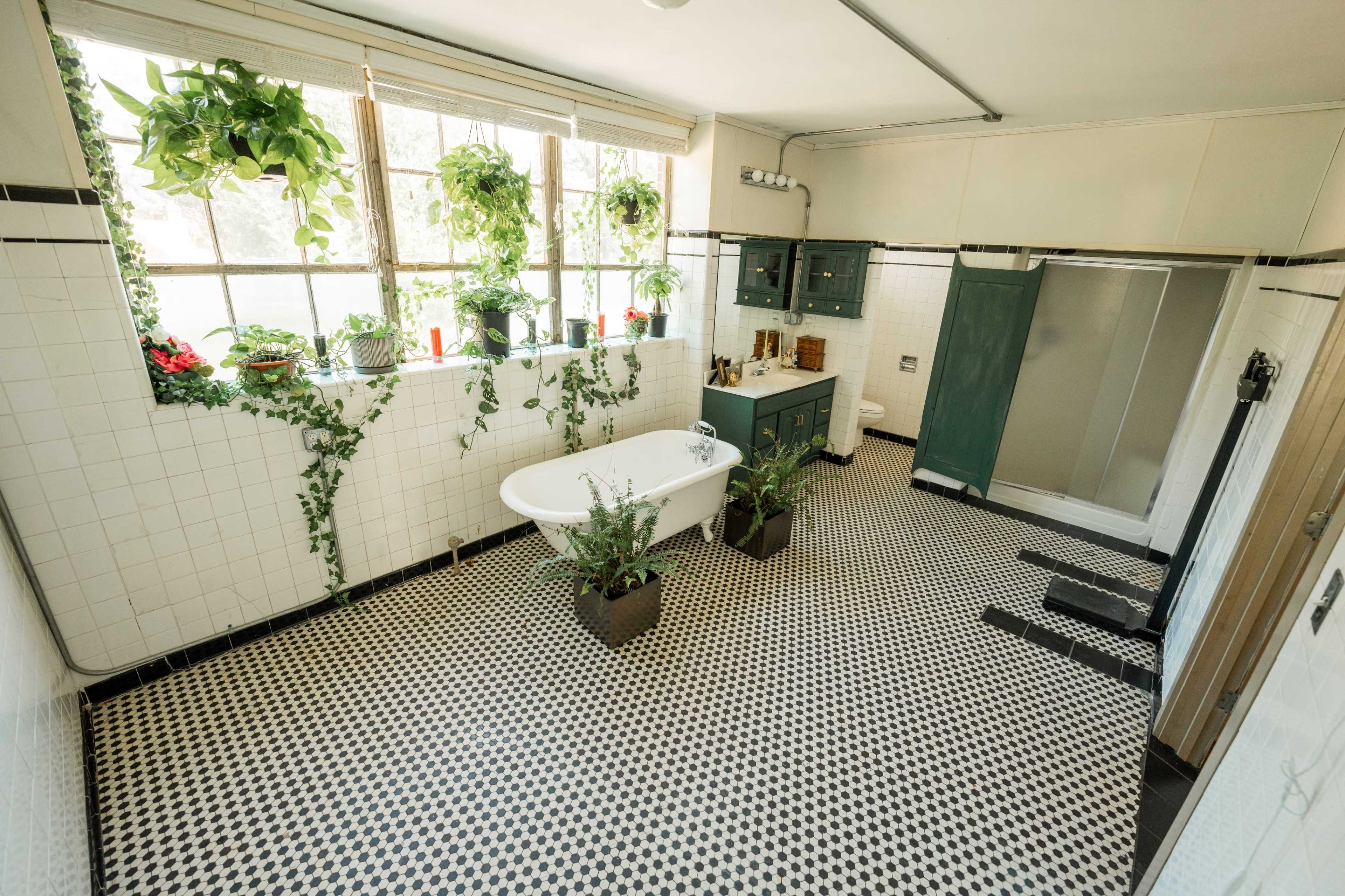 A spacious bathroom featuring a freestanding bathtub, green plants on windowsills, and a checkered black and white tiled floor.
