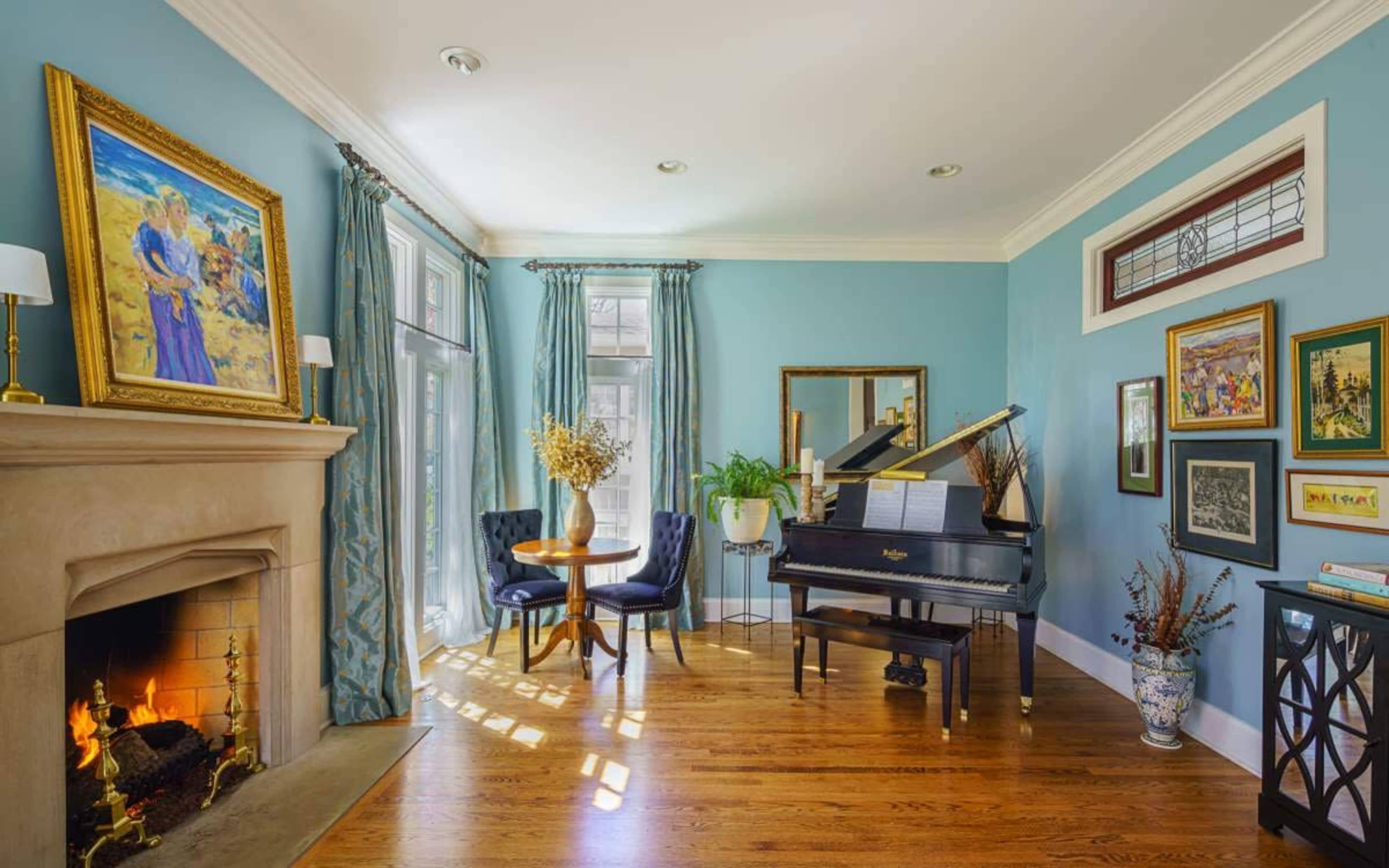The image features a cozy living room with a grand piano, a small table with chairs, a fireplace, and an array of framed artwork on light blue walls.