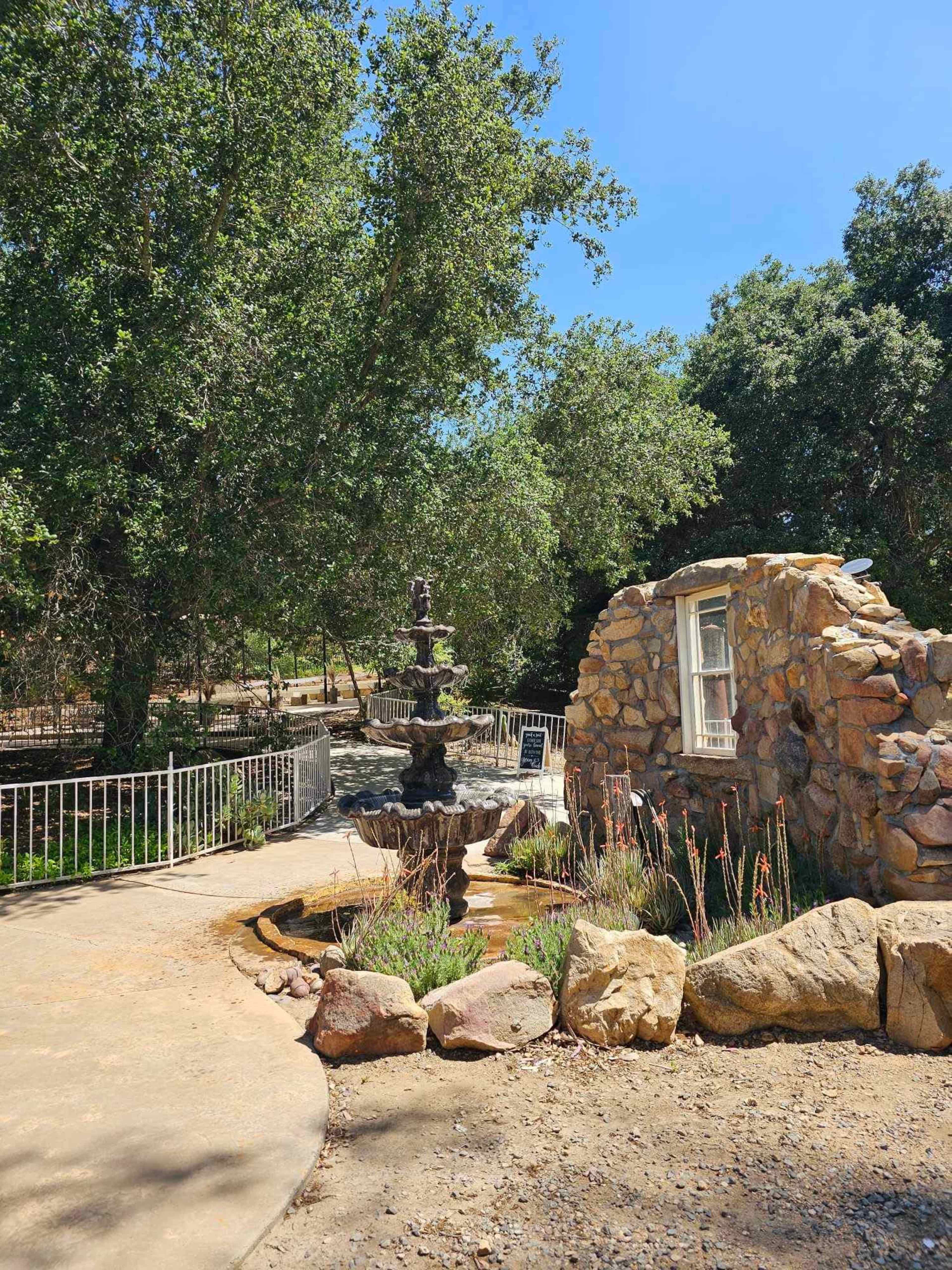 A stone fountain surrounded by a landscaped area features a small building and trees in the background.