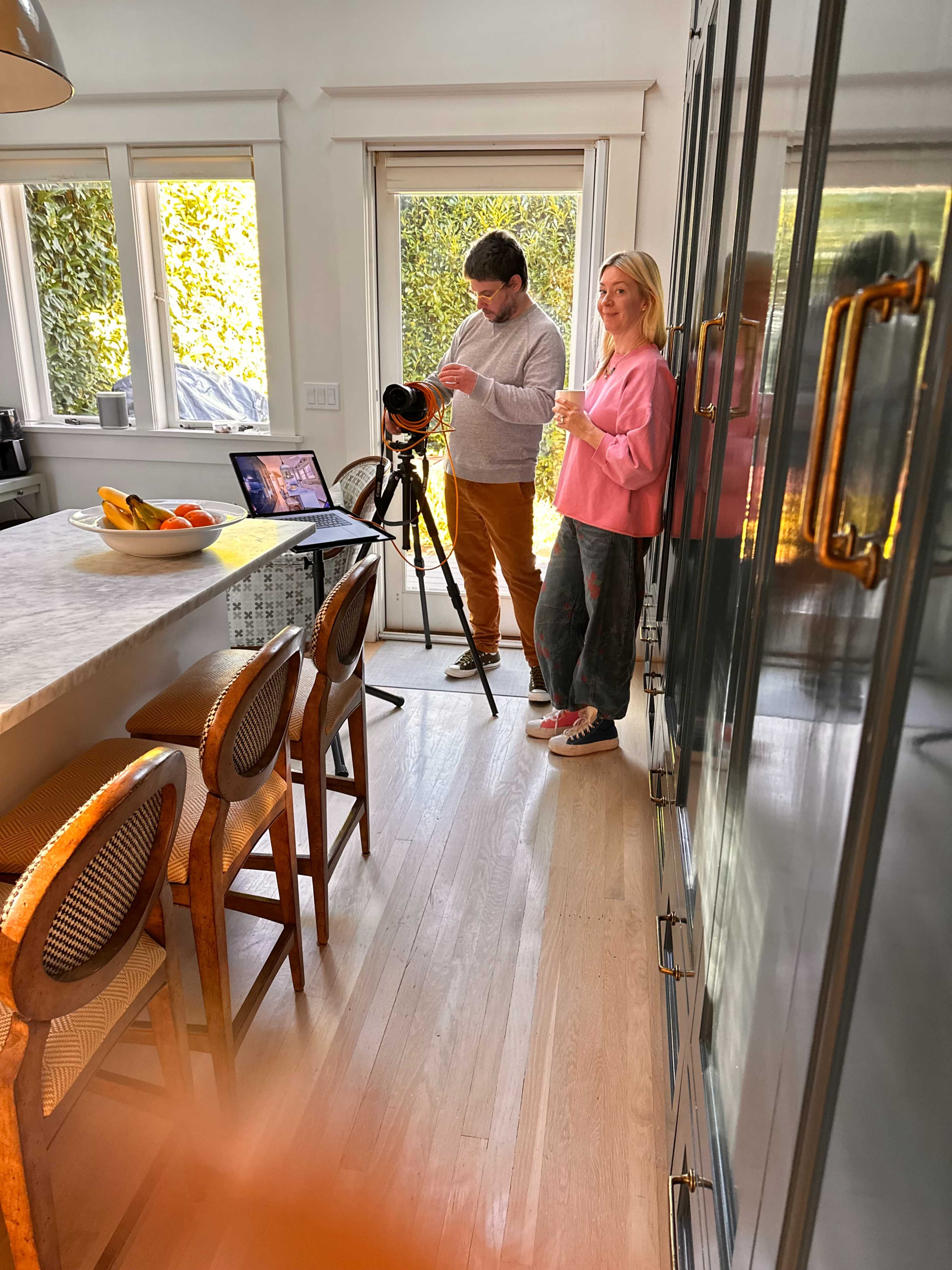 A person is setting up a camera on a tripod in a bright kitchen while another person stands nearby holding a drink.