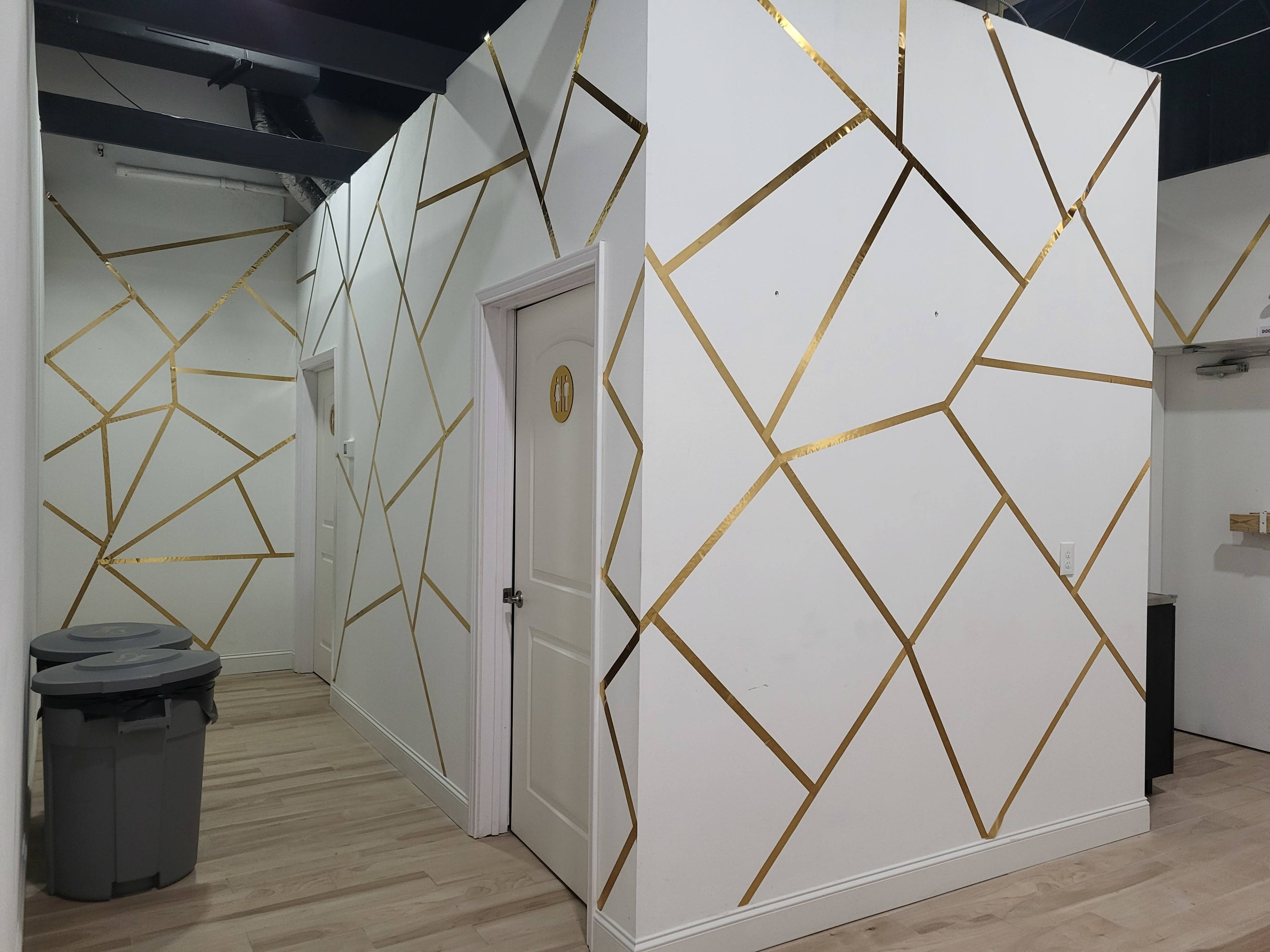 The image shows a hallway featuring white walls with a geometric gold foil design and a wooden floor, along with two gray trash bins on the left side.