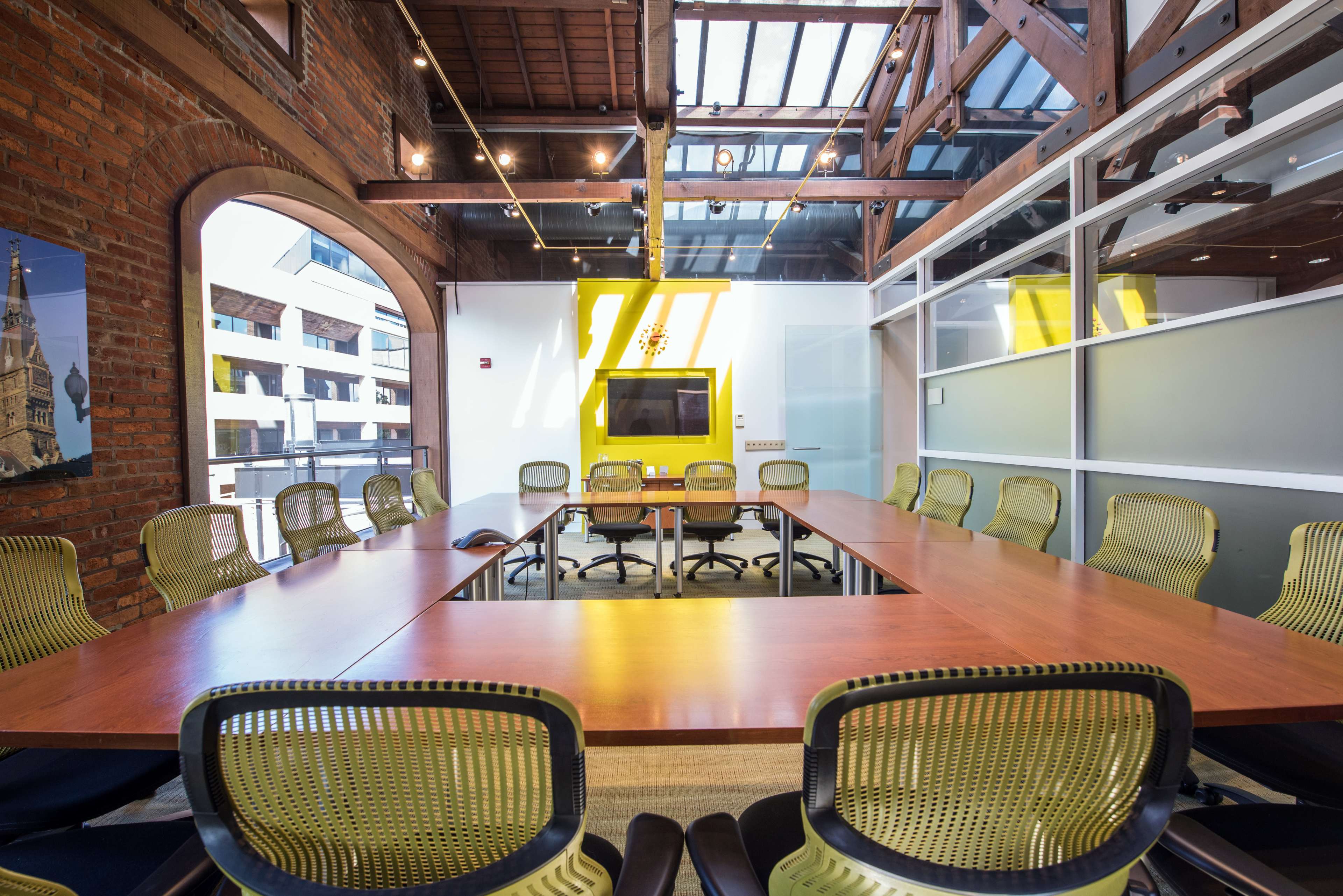A large conference room features a rectangular wooden table surrounded by green ergonomic chairs, with exposed brick walls and skylights above.