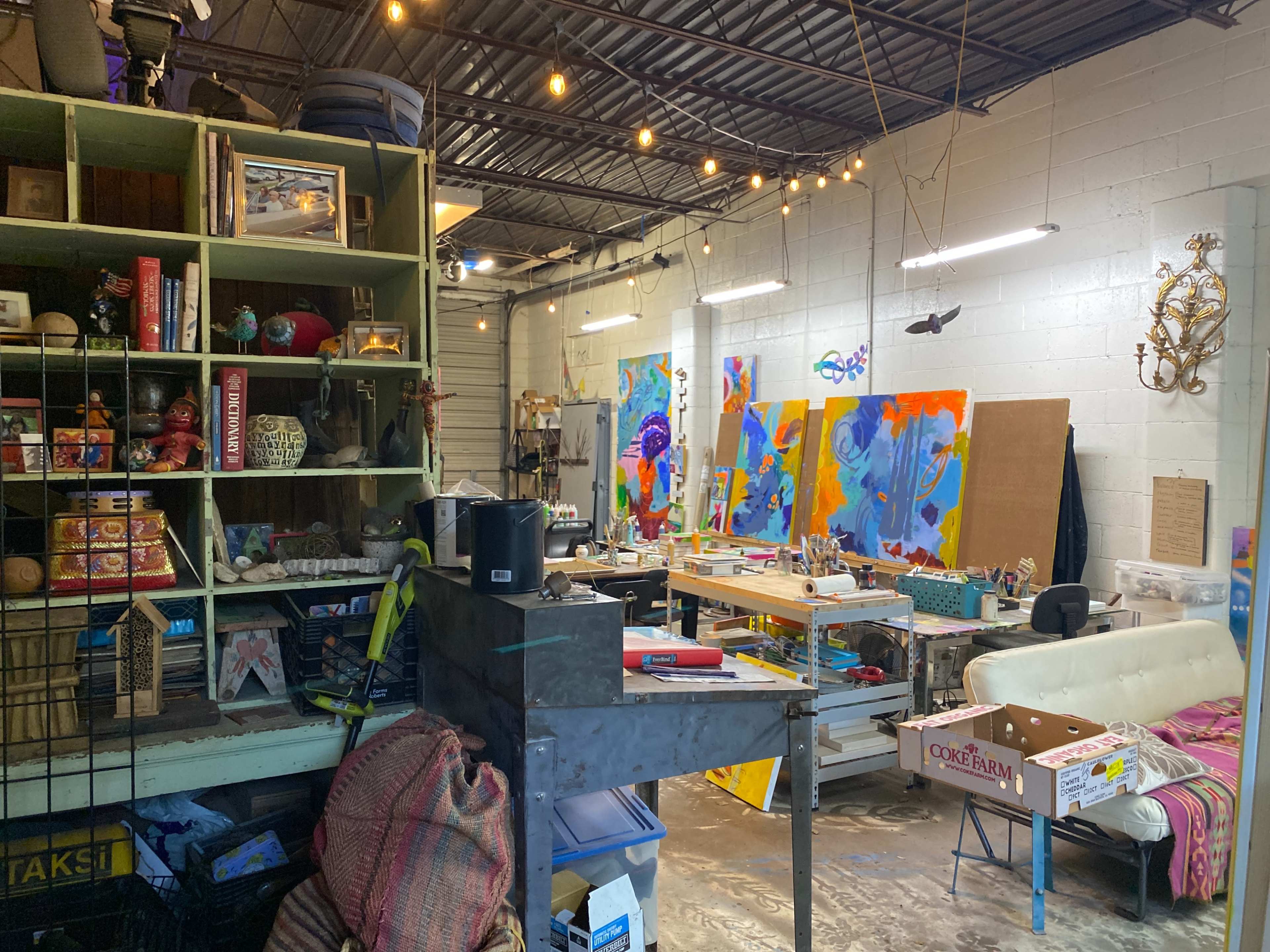 The image shows an art studio filled with various supplies, furniture, and vibrant paintings on the walls.