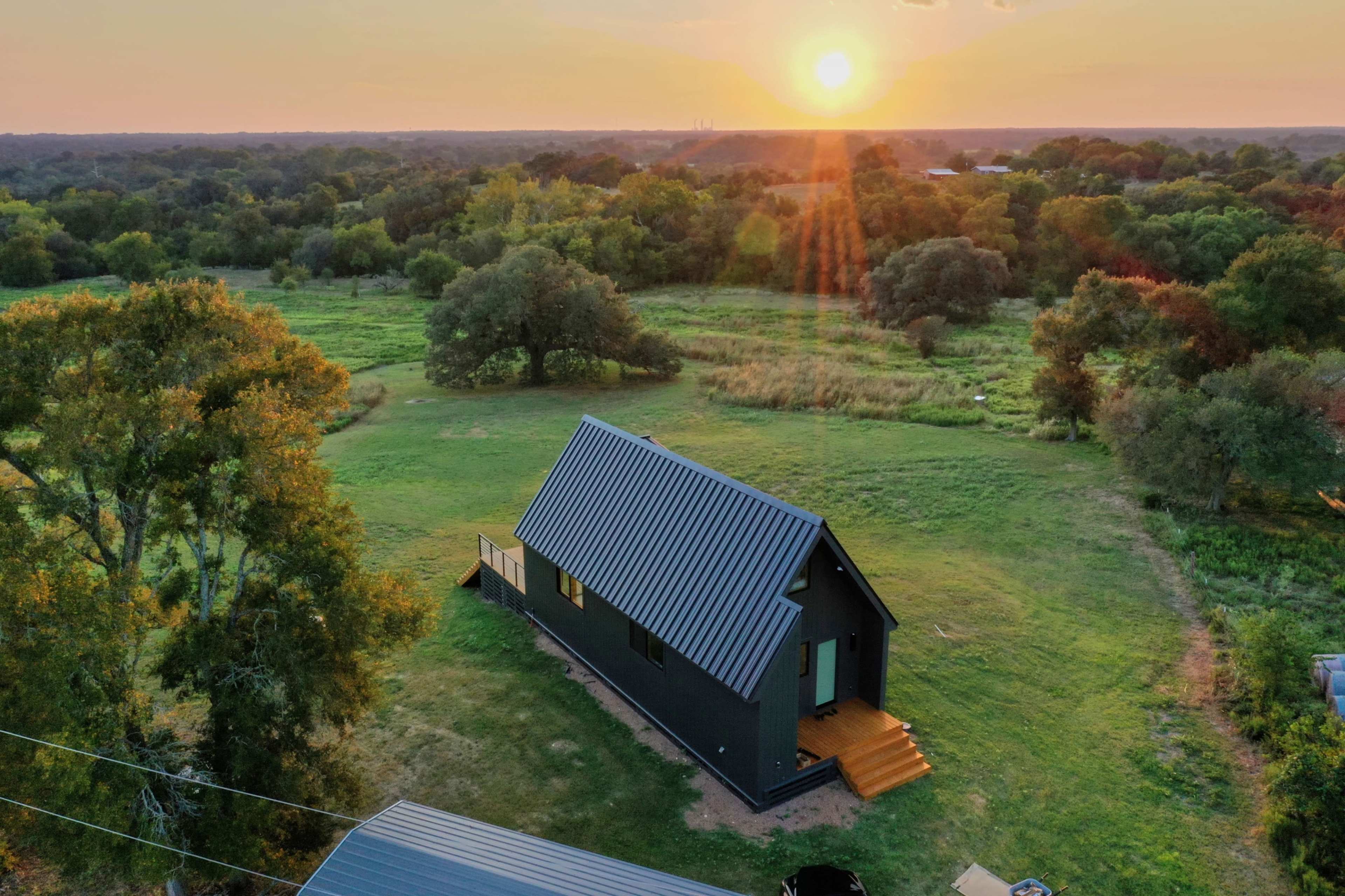 A black-roofed house sits in a green landscape with trees, as the sun sets in the background.