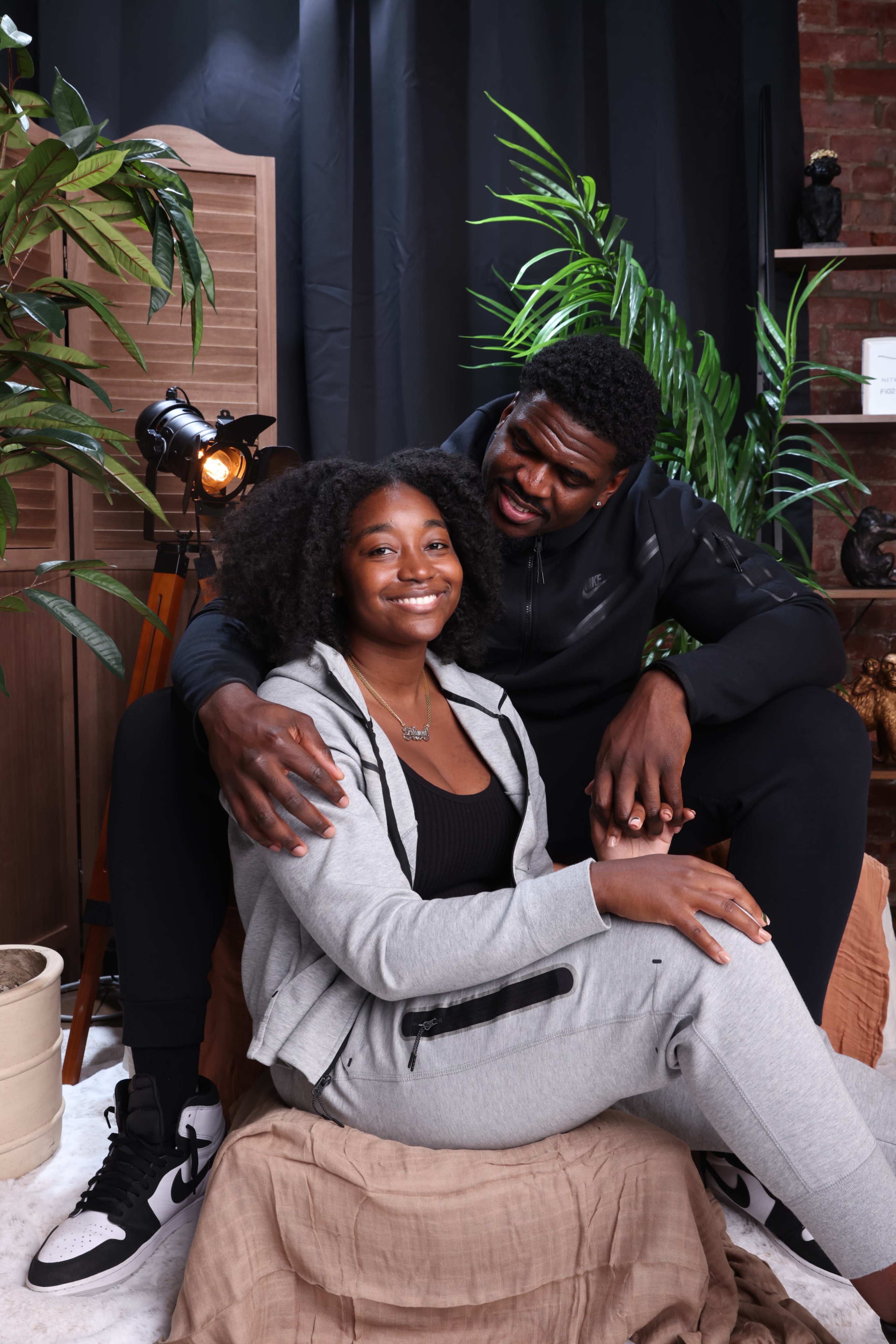 A man and woman sit together on a couch, surrounded by plants and decorative items, with the man playfully leaning towards the woman.